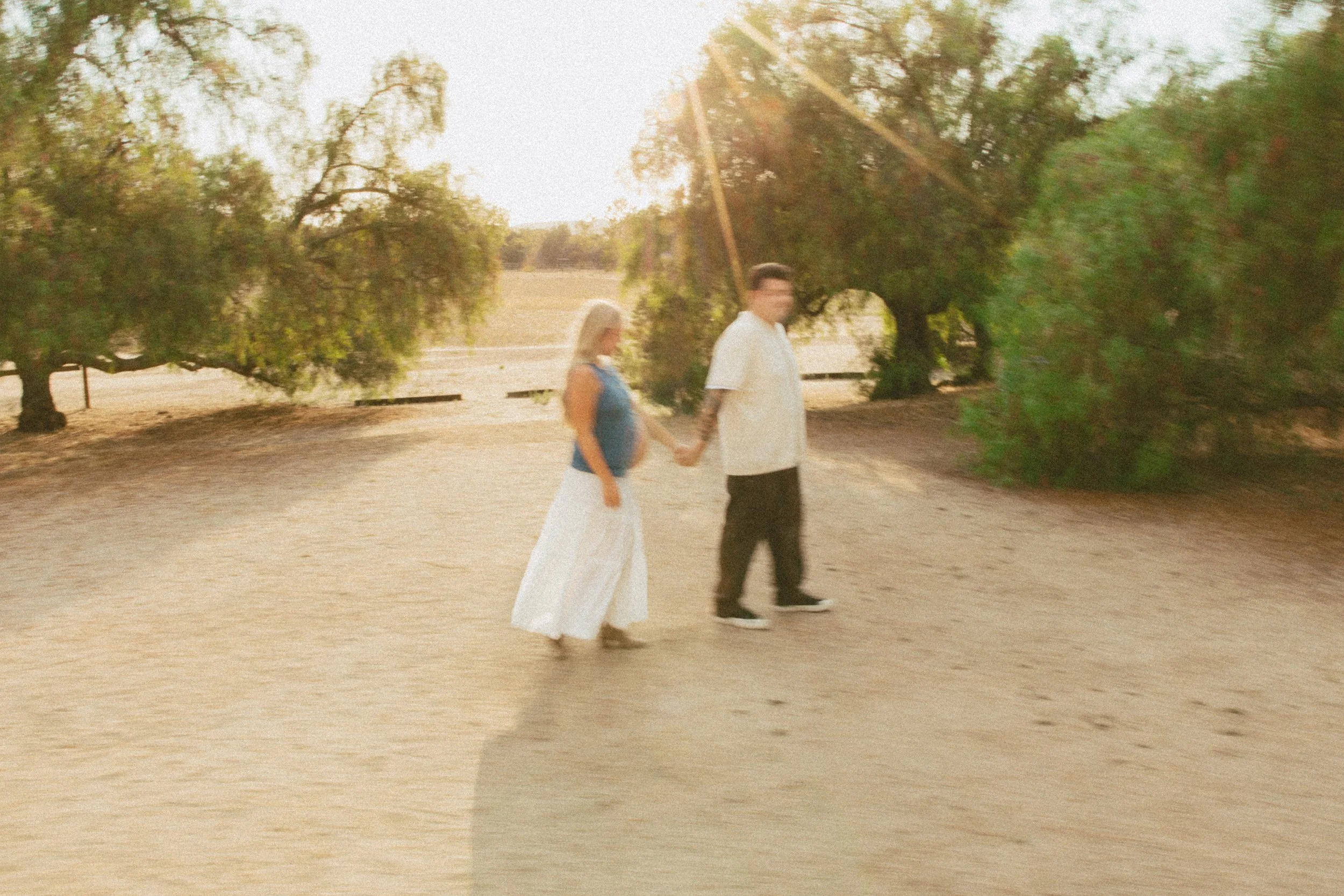 A couple holding hands walking outdoors on a sunny day surrounded by trees.