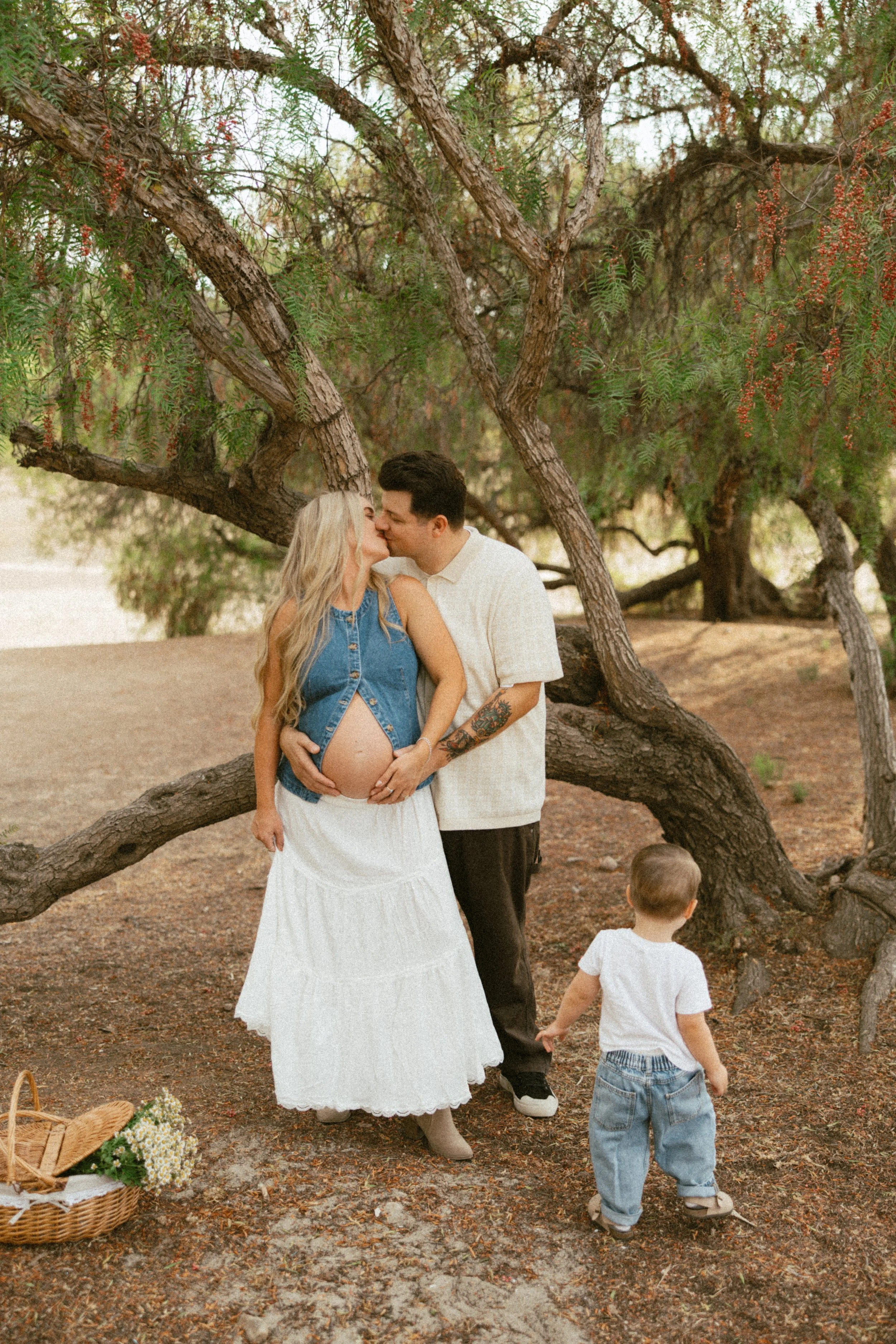 A couple sharing a kiss, with the woman pregnant, standing under a large tree in a park. Two small children are nearby, one holding the woman's hand, and a picnic basket with flowers is on the ground.
