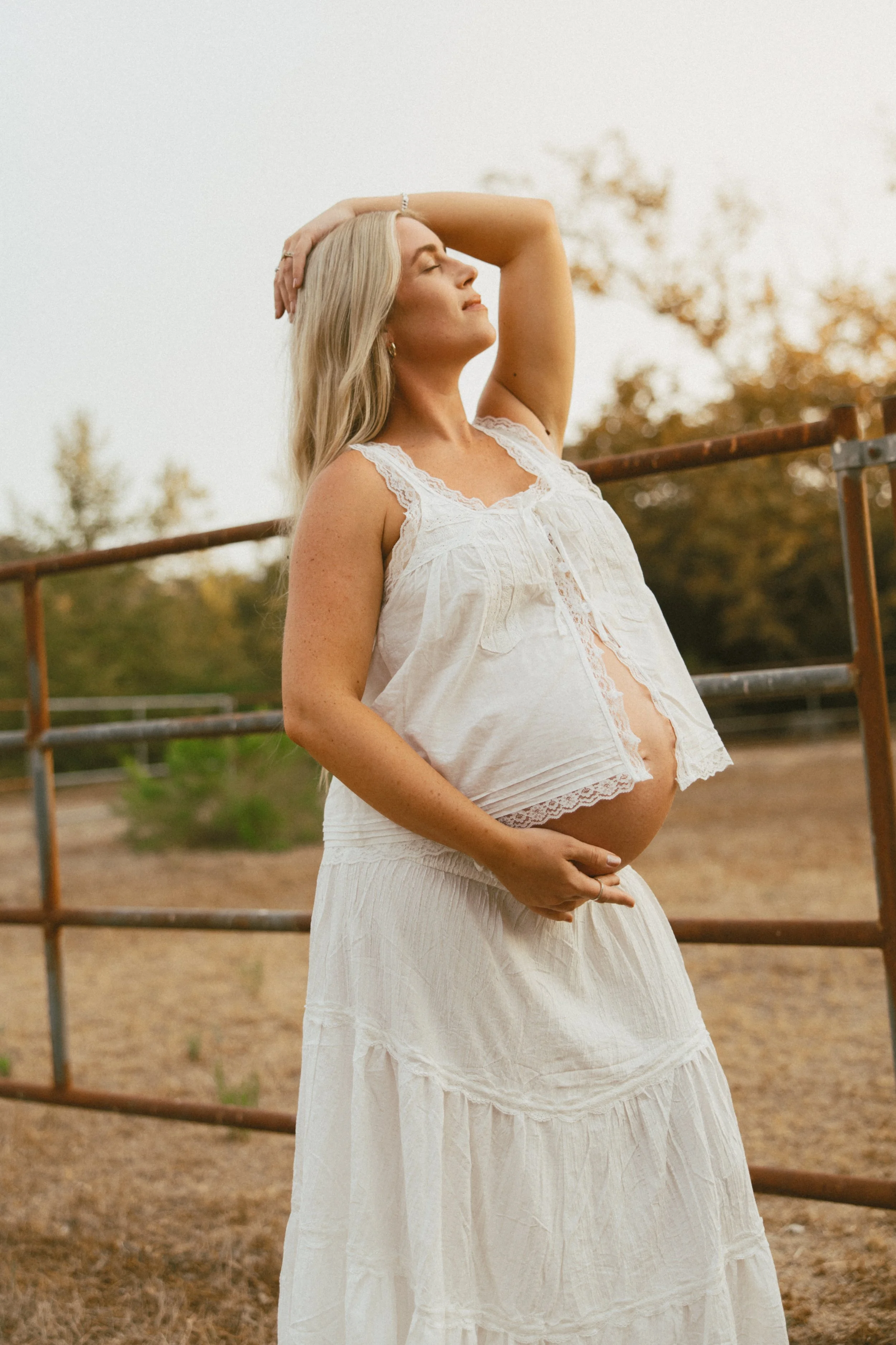 A pregnant woman in a white lace dress stands outdoors at sunset, with her eyes closed and hand on her head, gently cradling her belly.