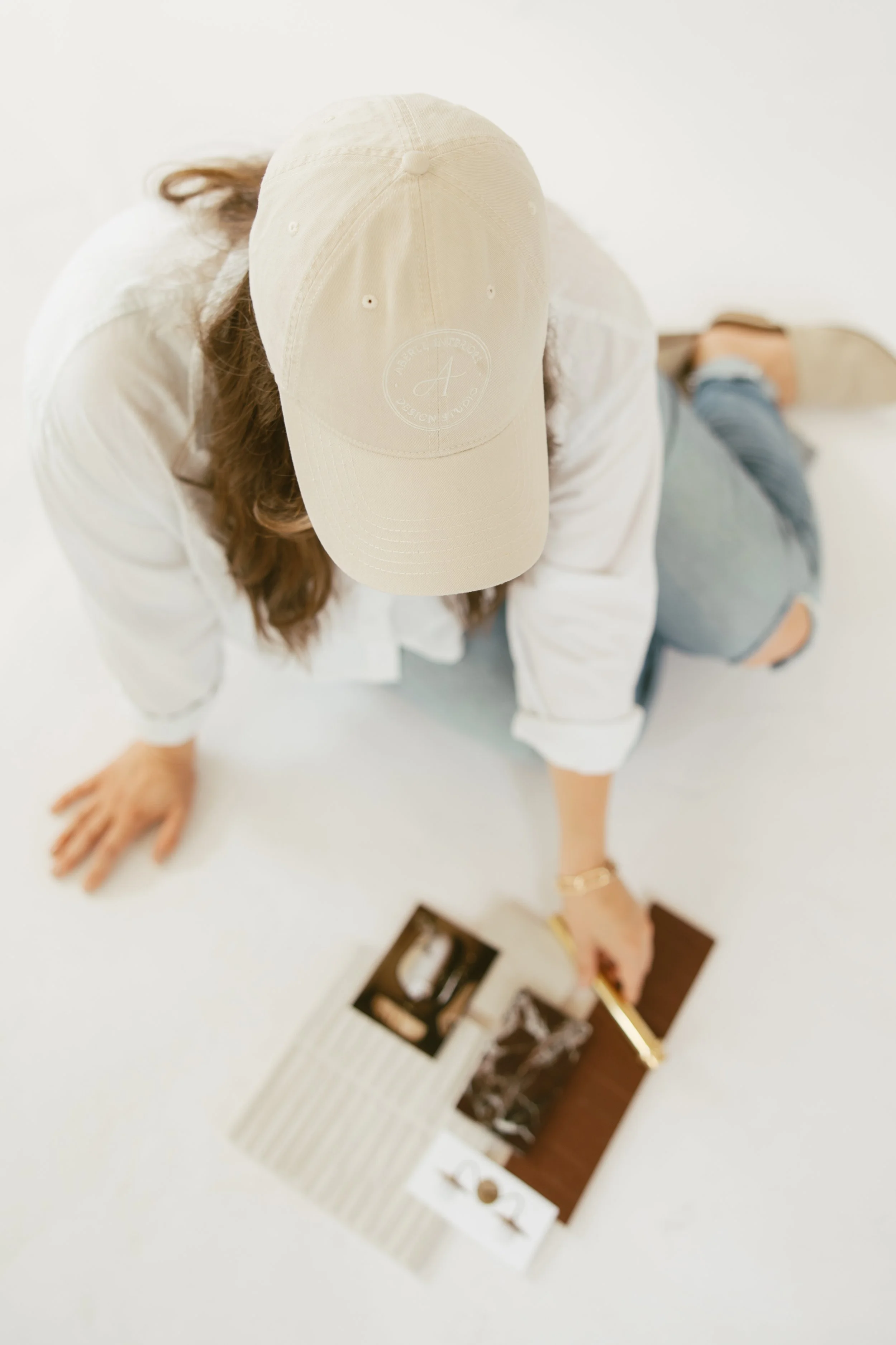 Top view of a person wearing a beige baseball cap, white shirt, and jeans, sitting on a white surface, with a magazine and a receipt nearby.