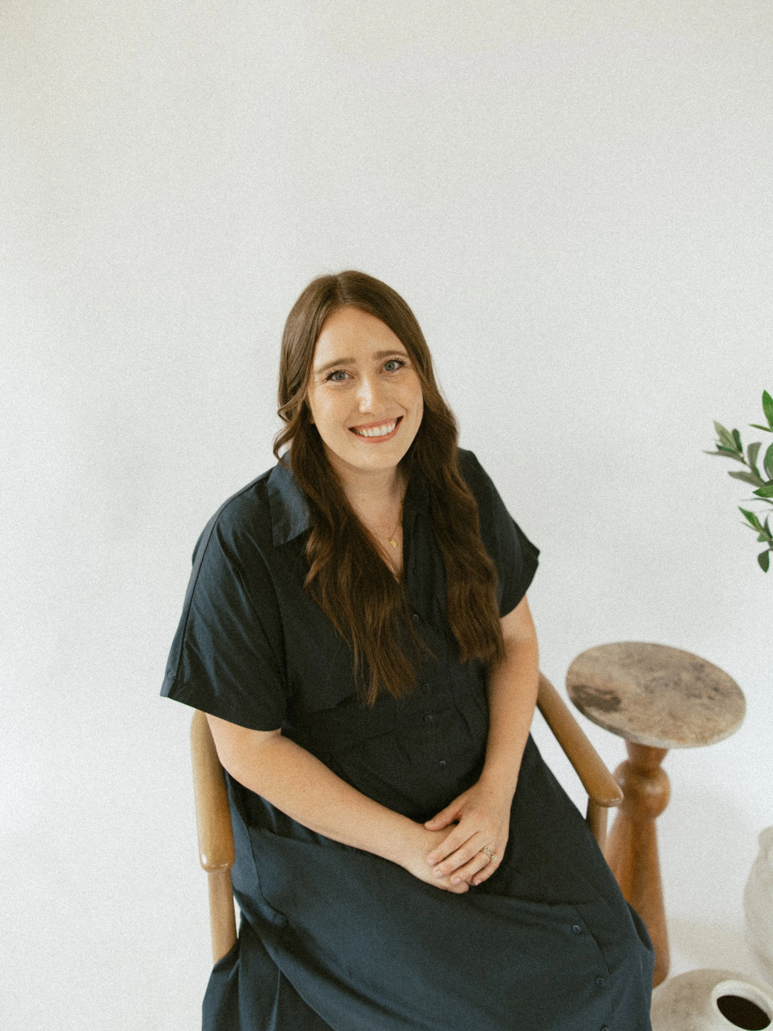 A woman with long brown hair and a black dress sitting on a wooden chair, smiling, with a JavaScript: All the GraphQL & React You Need to Know book on her lap. There is a small wooden table with a plant in a white pot nearby.