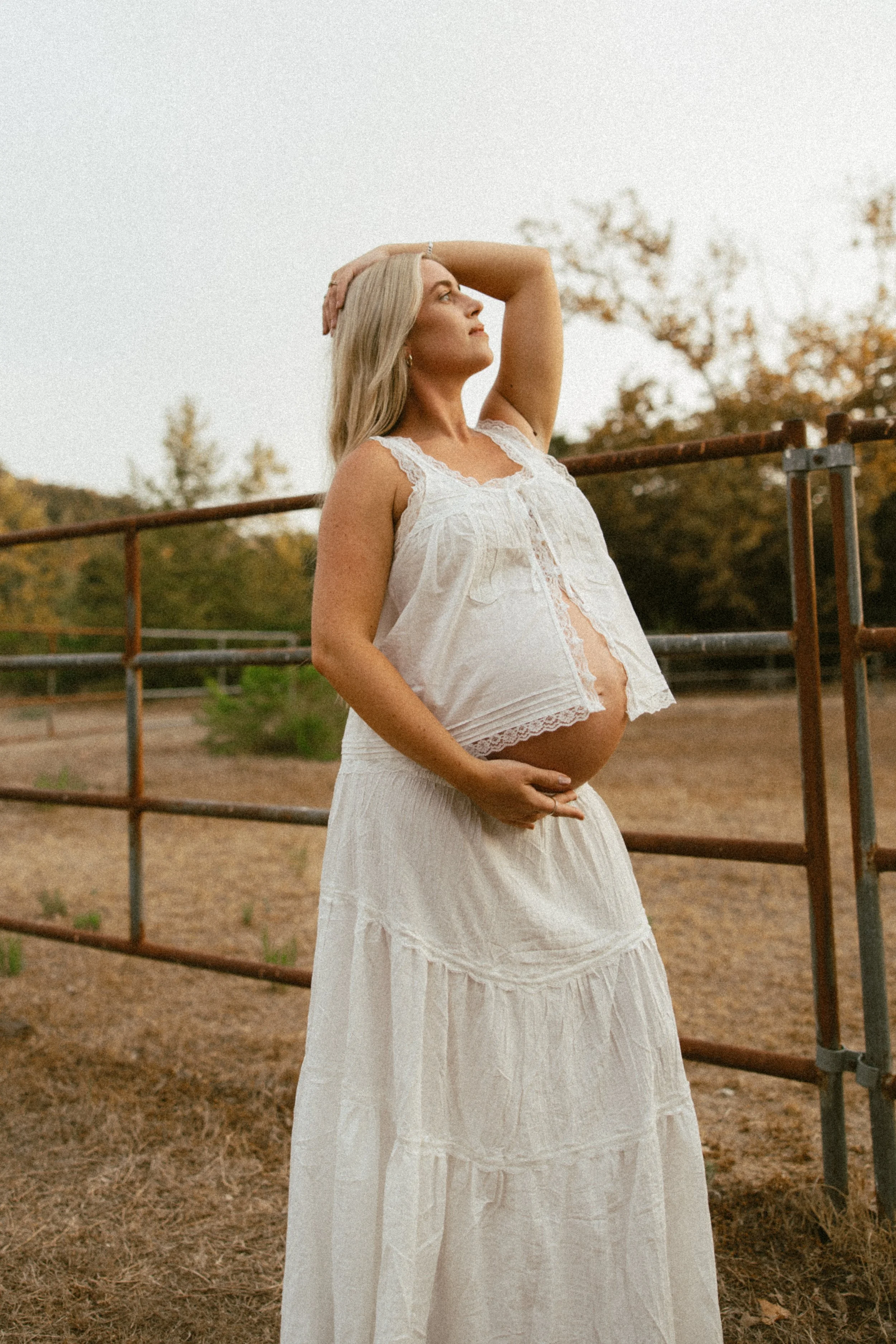 Pregnant woman with long blonde hair, wearing a white lace top and skirt, standing outdoors near a metal fence, with trees in the background during sunset.