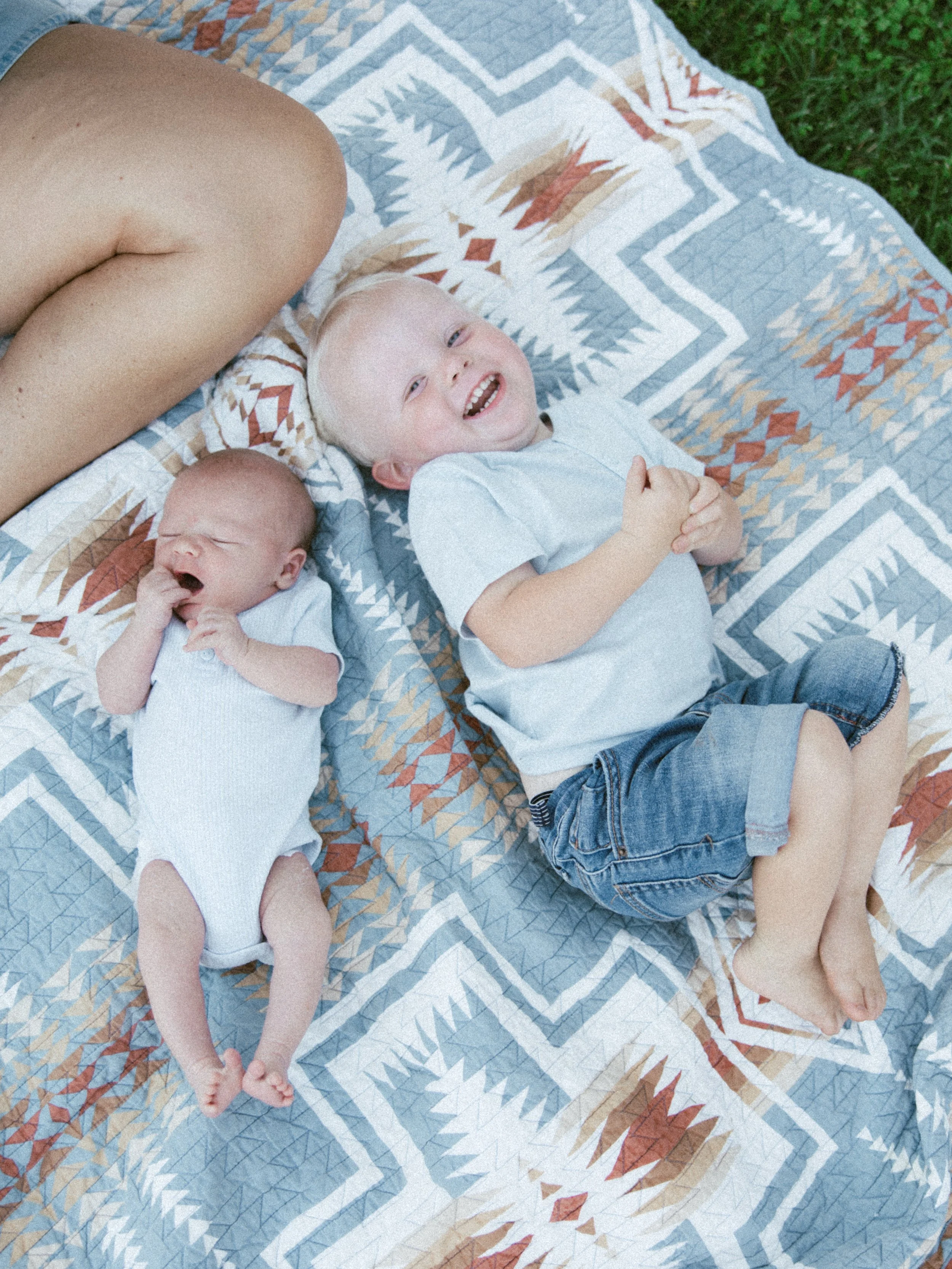 Two young children, a baby and a toddler, lying on a quilted blanket outdoors on grass. The baby is yawning and the toddler is smiling happily.