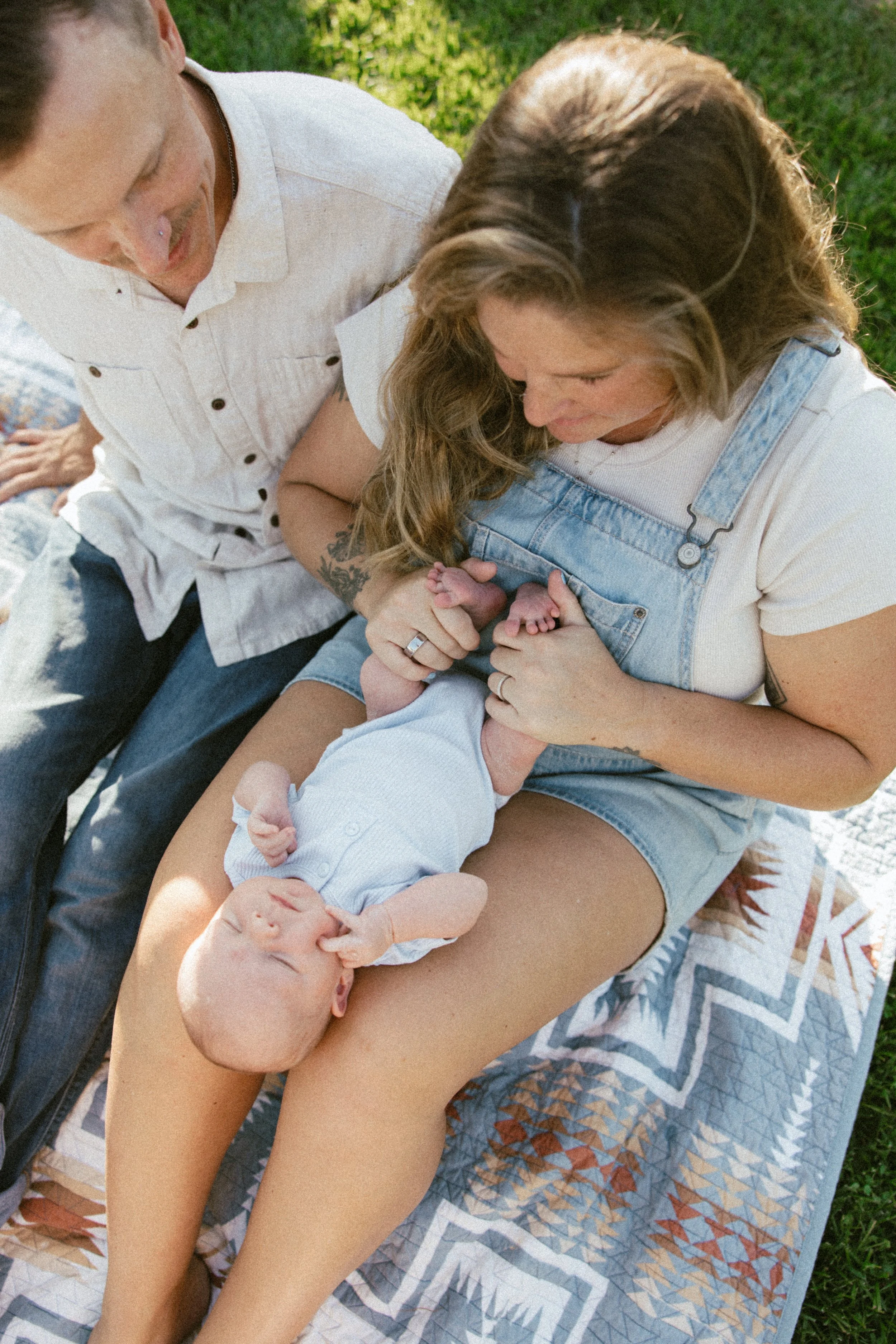 A woman with light brown hair, wearing overalls, sitting outdoors on a blanket on the grass, holding a newborn baby with a bald head and light skin, while a man with light brown hair, wearing a white shirt, looks on.