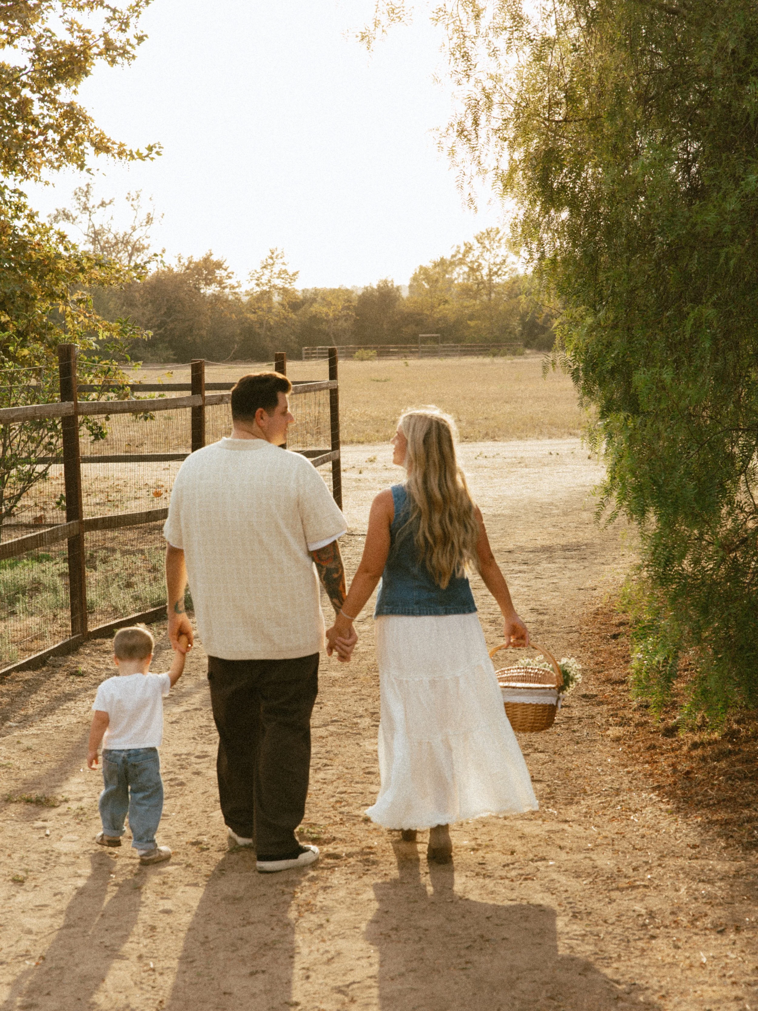 A couple holding hands and walking outdoors on a sunny day, with a young child holding one of the woman's hands. The woman is wearing a long white skirt and a denim vest, the man is dressed in dark pants and a light shirt, and they are carrying a pic