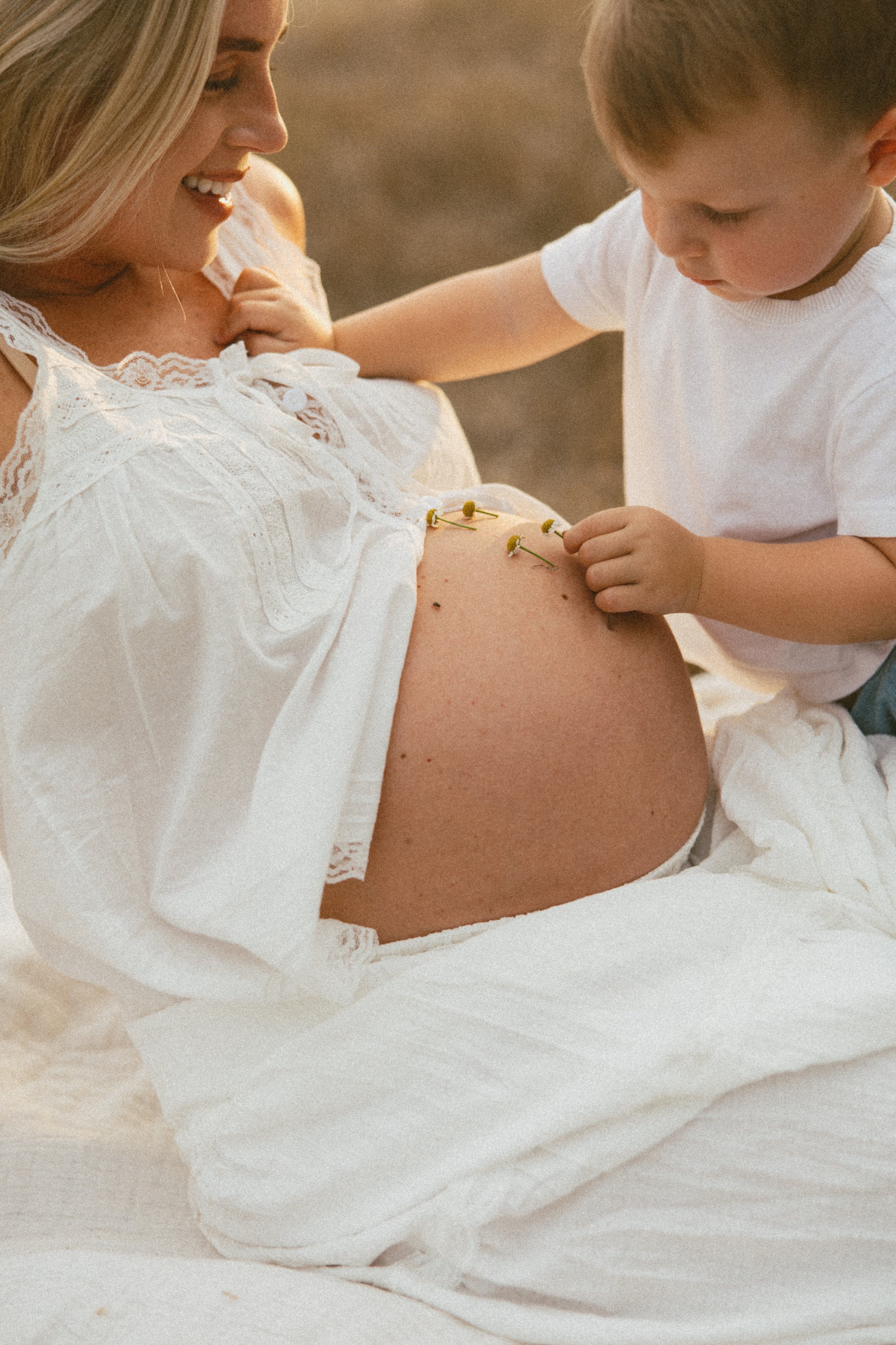 A pregnant woman smiling while looking down, and a young boy touching her pregnant belly with daisies on her stomach, both wearing white clothes.