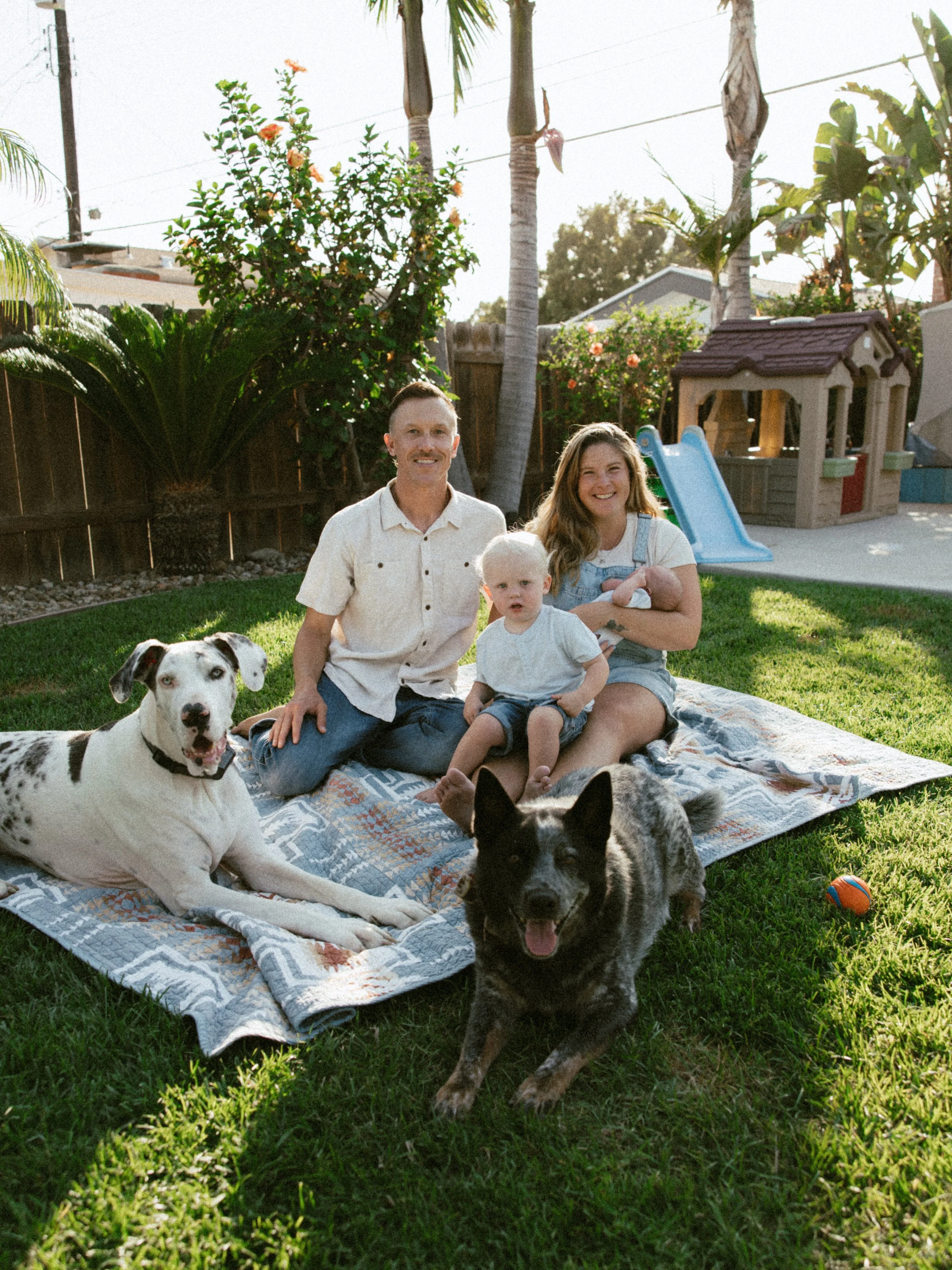 A family of three sitting on a blanket in a backyard with dogs, trees, and a playhouse, smiling and enjoying the sunny day.