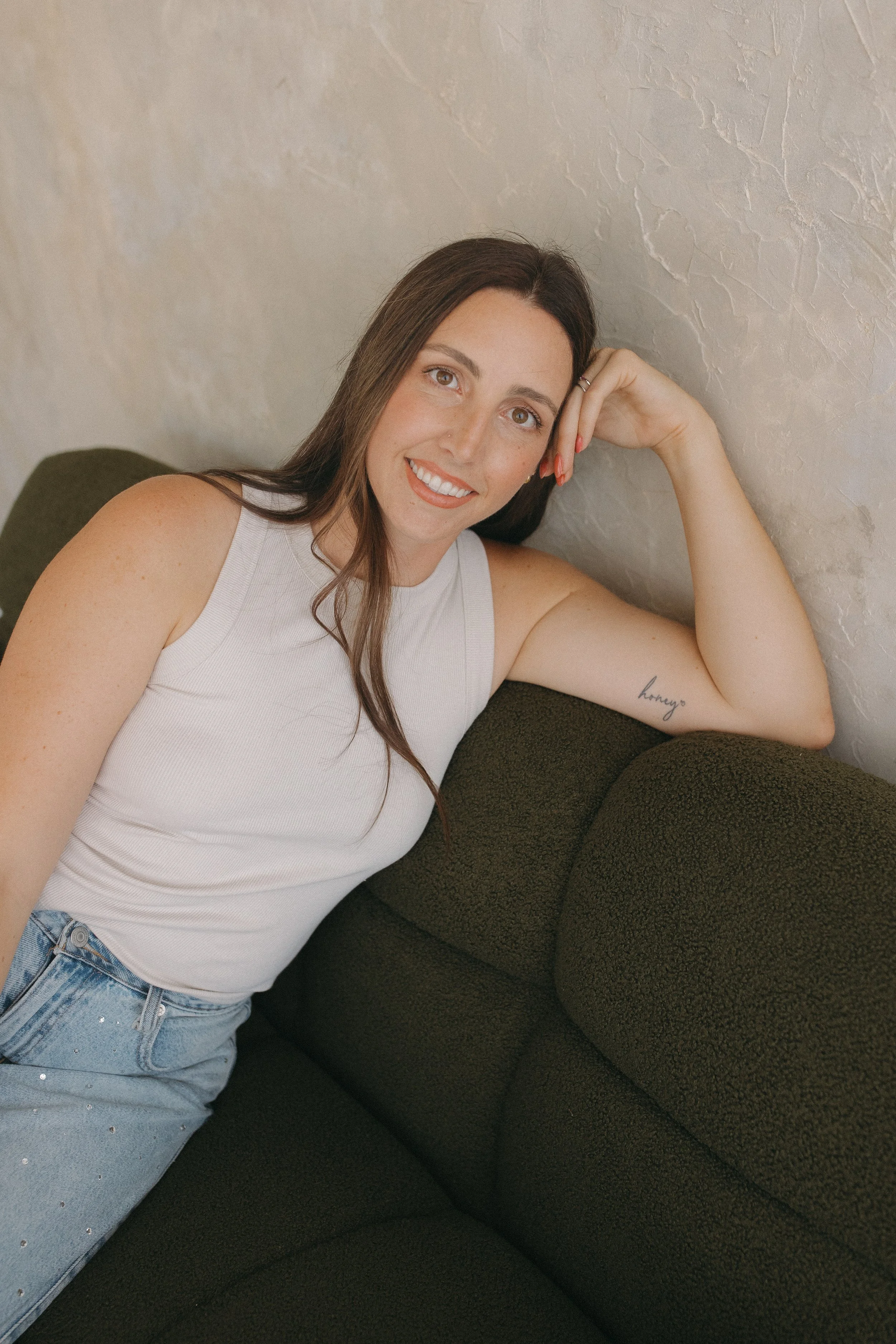 A woman with long brown hair and light skin smiling while lying on a dark green couch, resting her head on her hand, wearing a white sleeveless top and light blue jeans with embellishments.