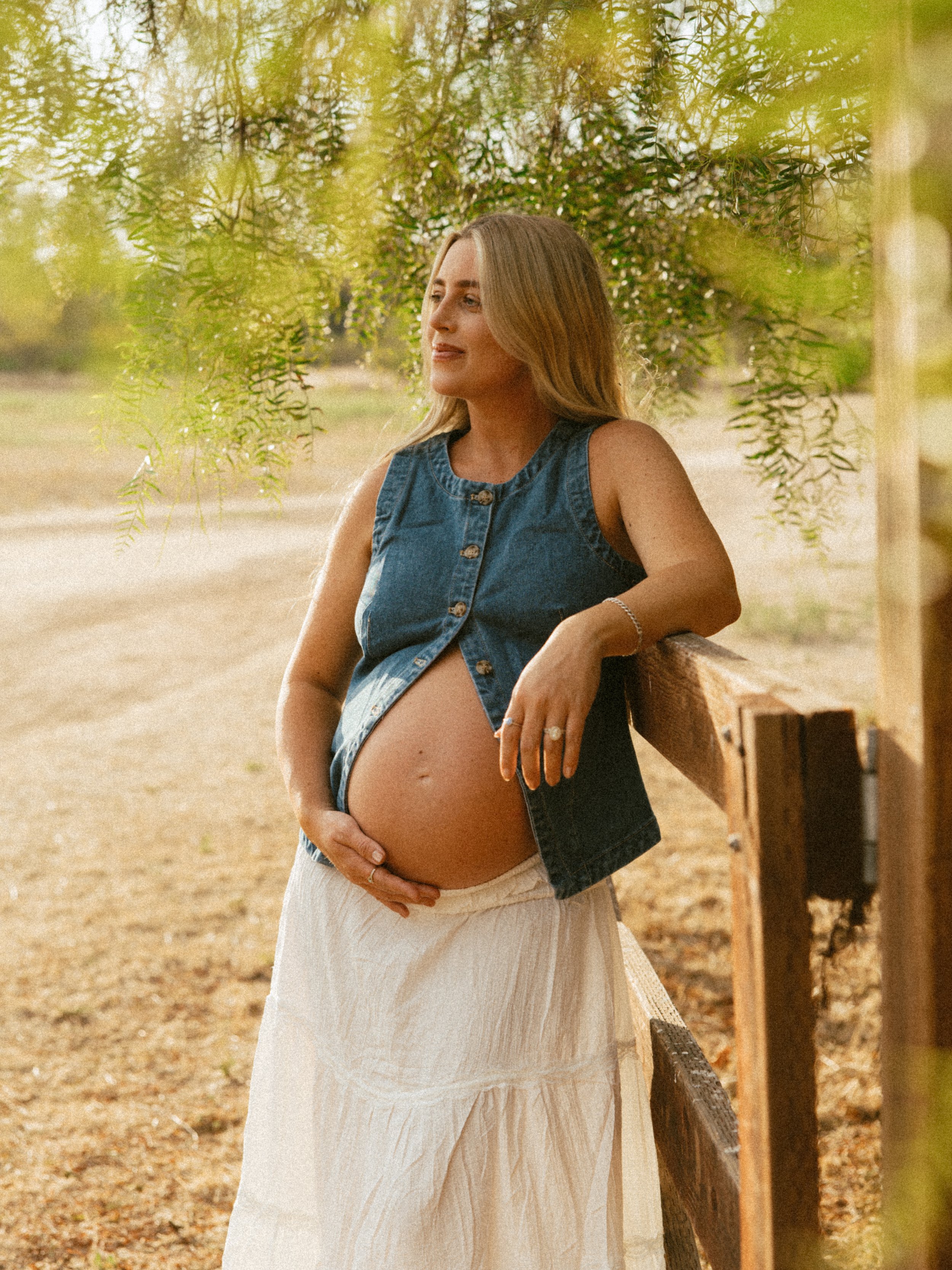 A pregnant woman with long blonde hair standing outdoors near a wooden fence, holding her belly and looking off to the side, surrounded by green foliage.