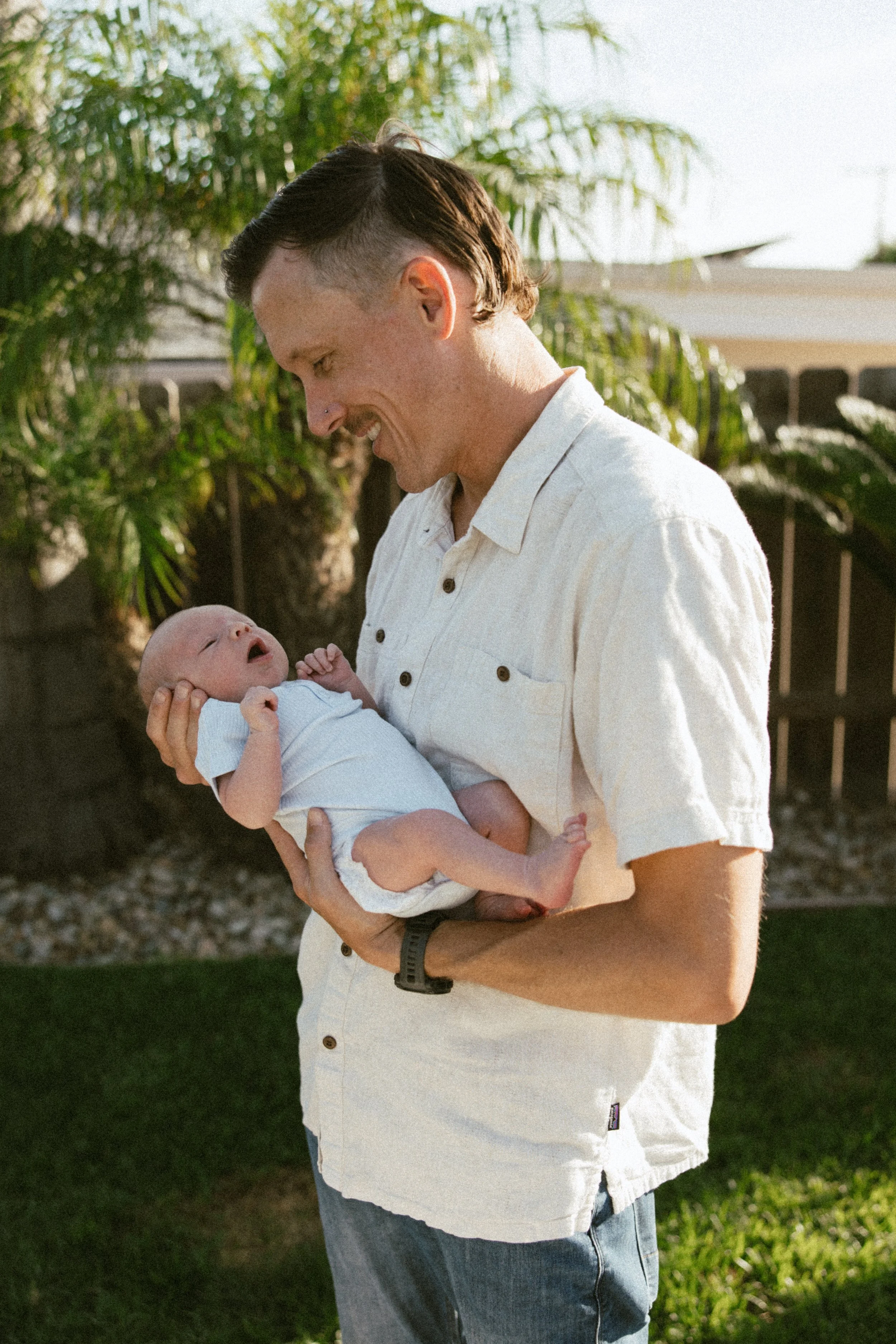 A man holding a baby outdoors with a garden and trees in the background.