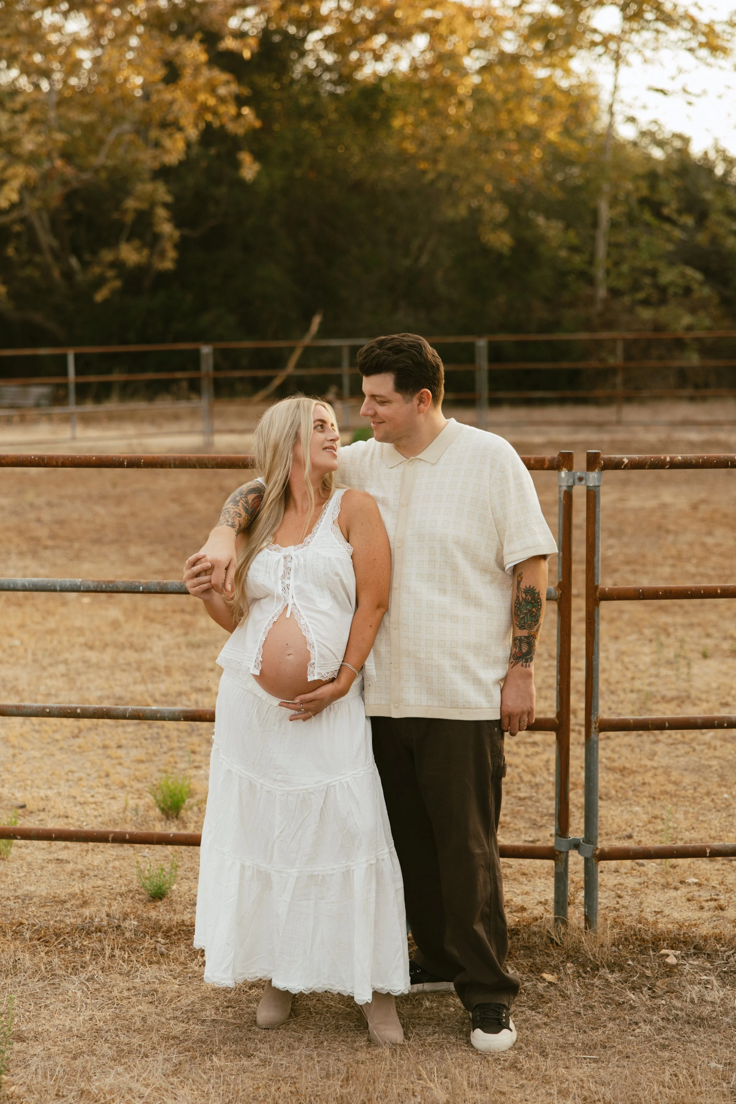A pregnant woman in a white dress and gray boots standing next to a man in a beige shirt and black pants, both smiling and looking at each other, in an outdoor setting with trees and a metal fence in the background.