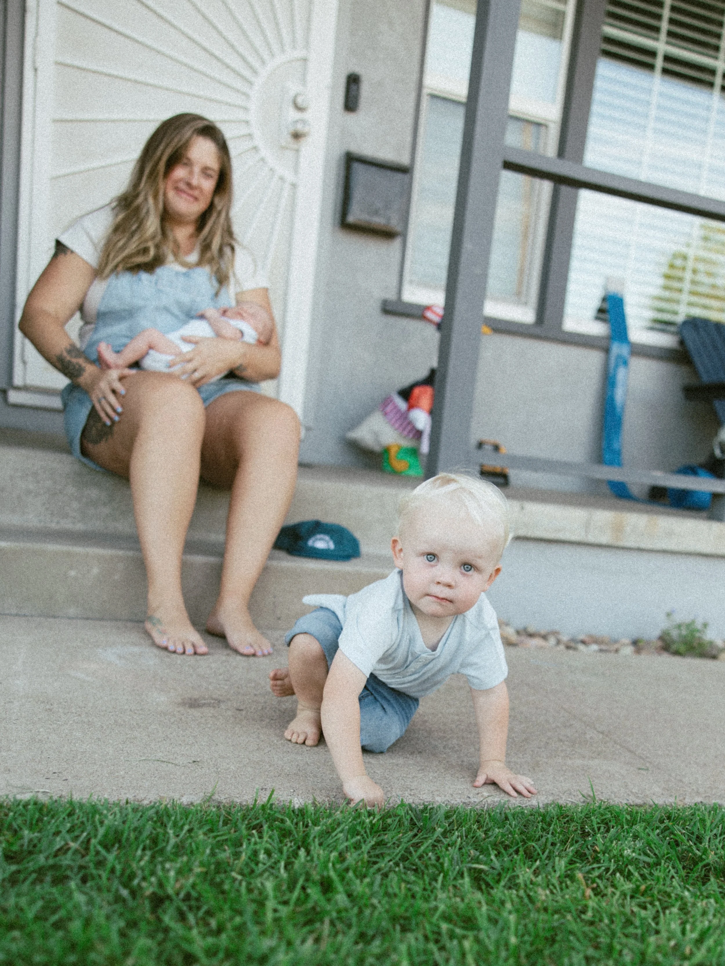 A woman sitting on front porch steps holding a baby, with a young boy crawling on the ground in front of her and looking up at the camera. The porch has a white door and gray railings, with toys and house items around.