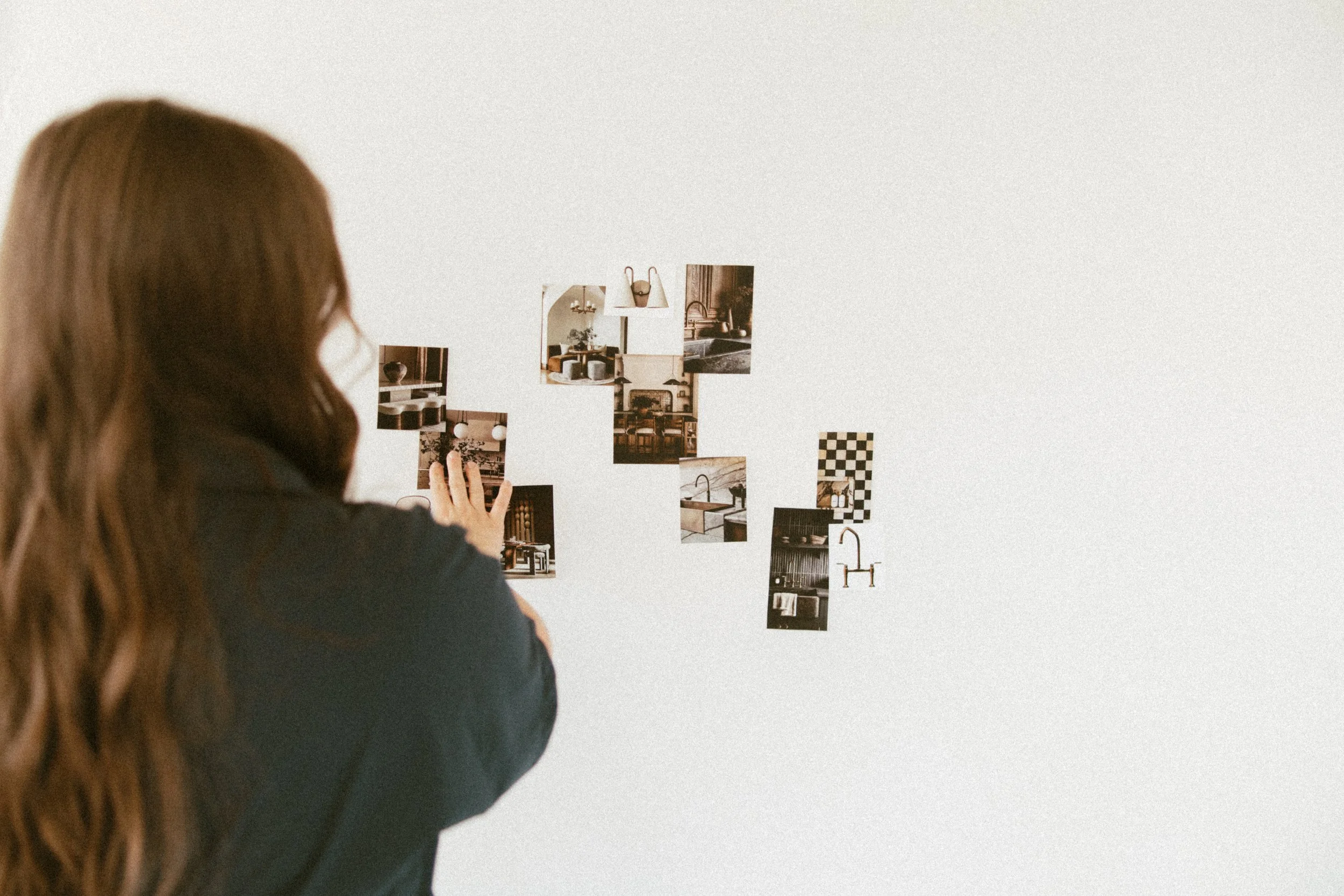 A person with long red hair and glasses viewed from behind, adjusting or placing photographs or images on a white wall.