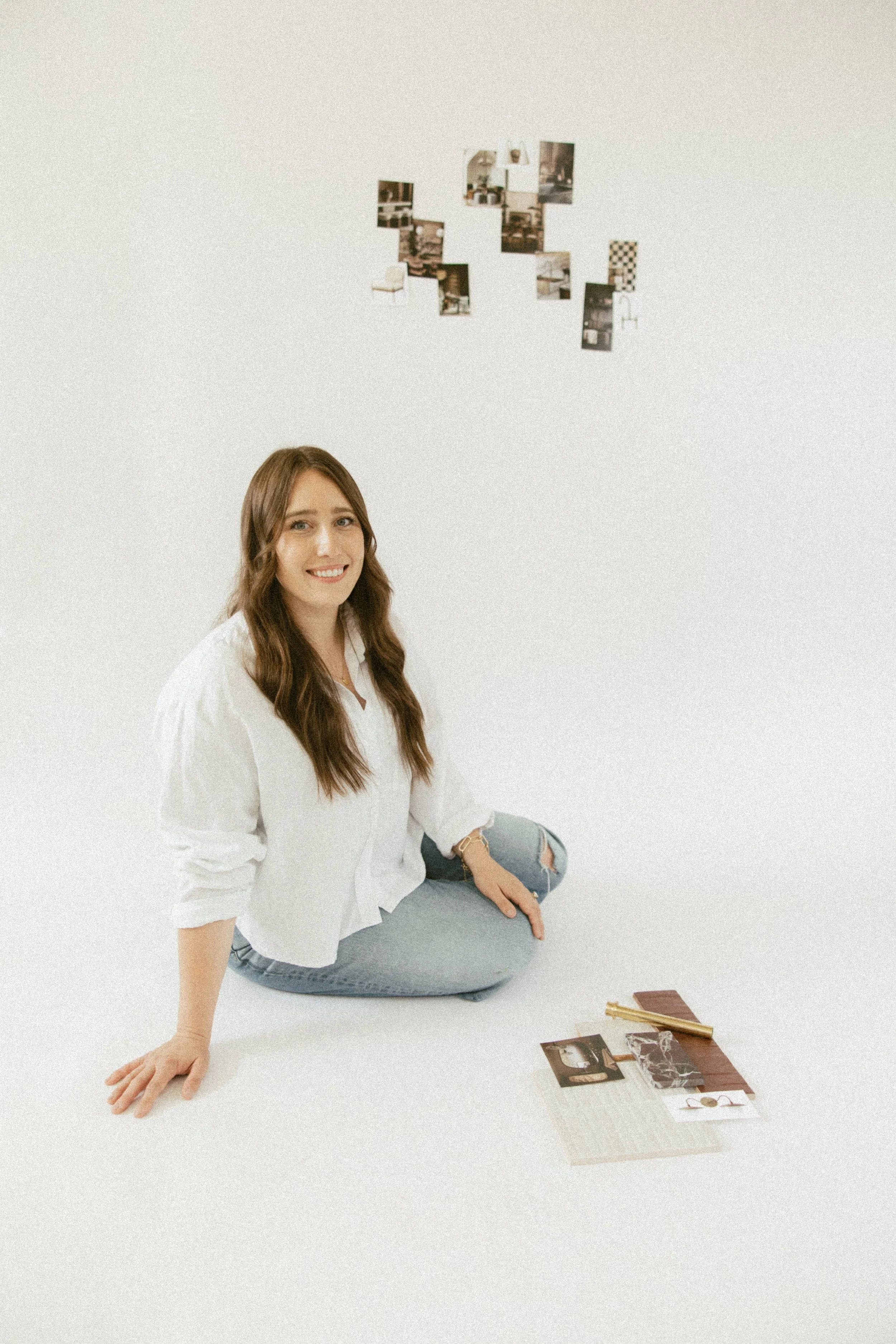 A young woman with long brown hair, wearing a white shirt and ripped jeans, sitting on the floor, smiling at the camera. In front of her are various photographs, magazines, and a gold pen on a small brown notebook.