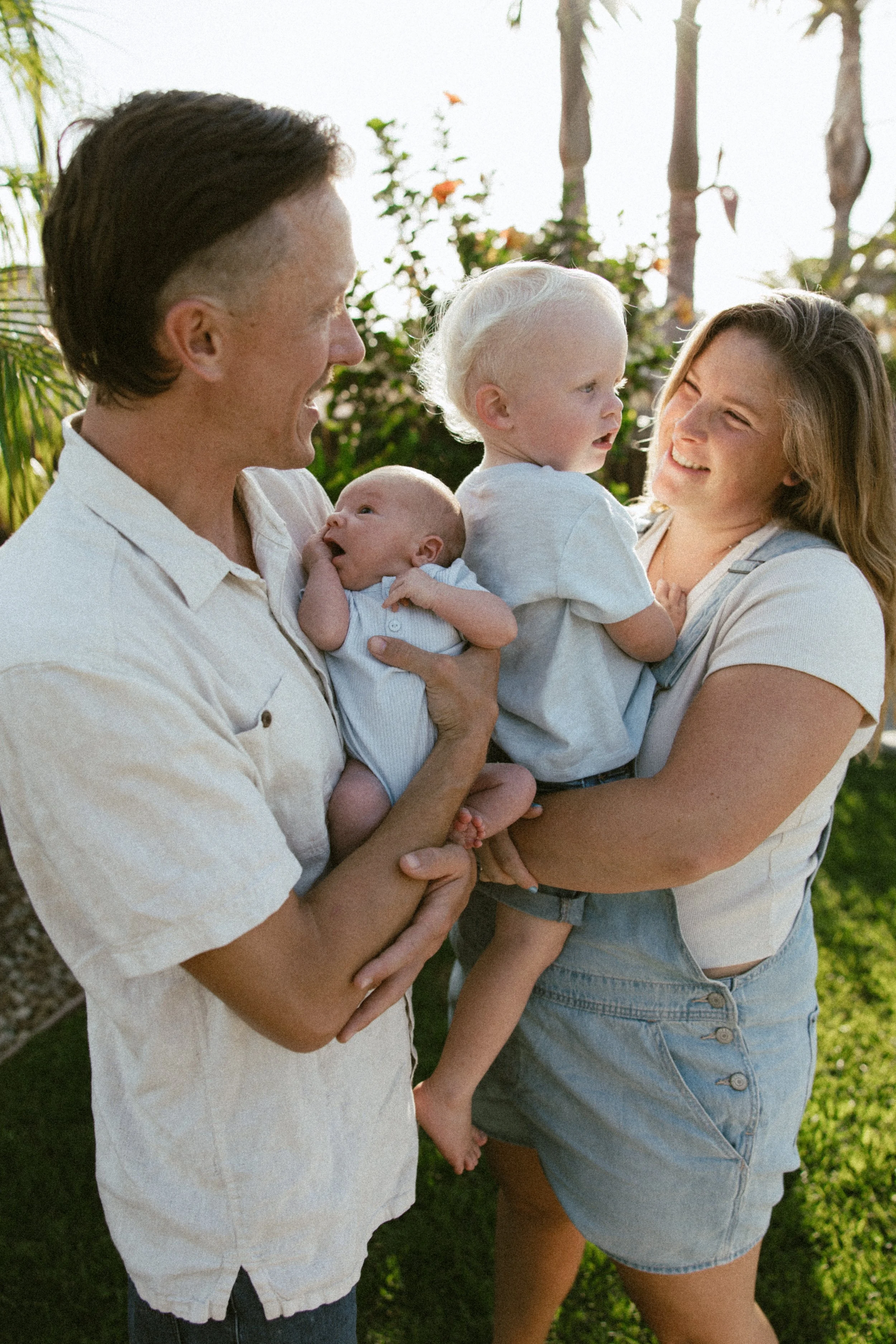 Family of four, parents with two young children, smiling and enjoying outdoors in a backyard with greenery and trees.