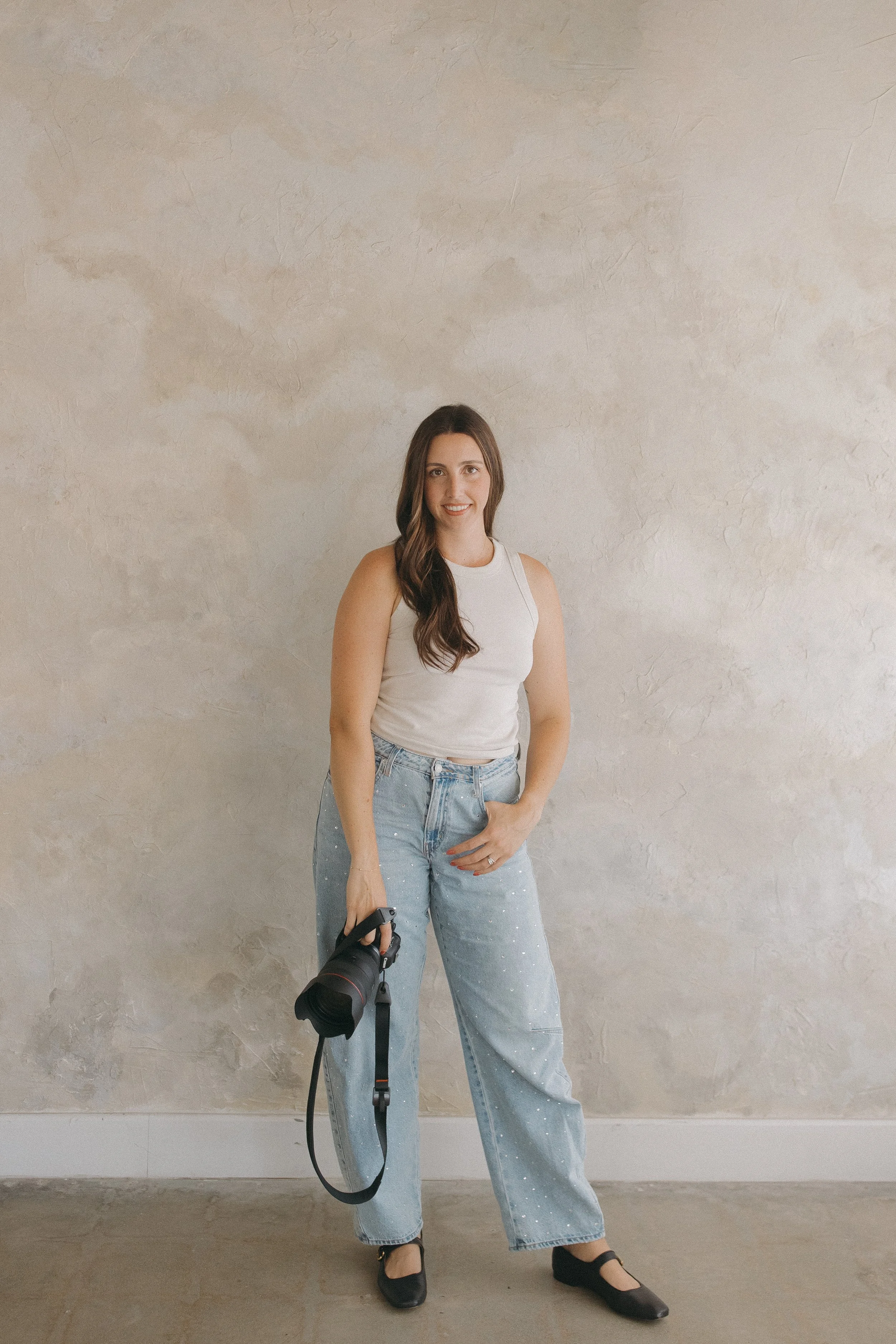 A woman with long brown hair wearing a white sleeveless top, light blue jeans with white specks, and black shoes, holding a camera with a strap in her right hand, standing against a beige textured wall.