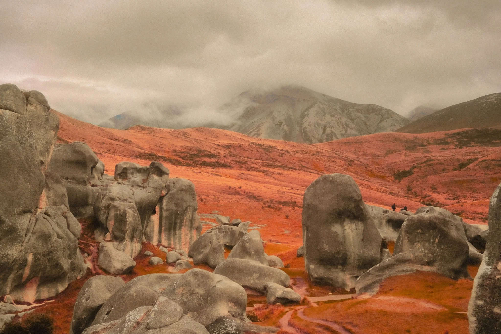 A landscape photo of rounded rocks in an orange-red valley with rolling hills and a cloudy sky, mountain peaks in the background.