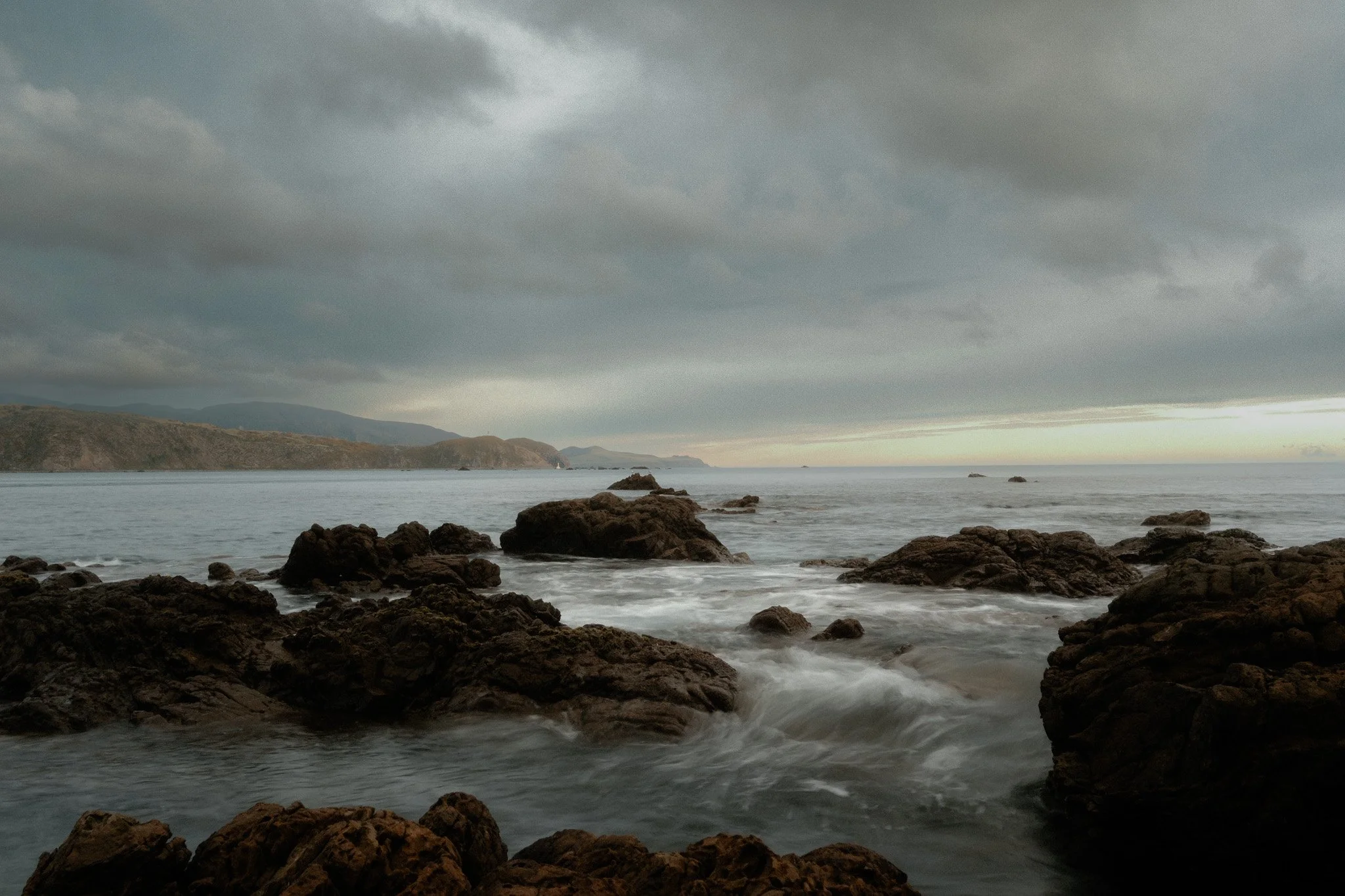 Overcast sky over a rocky shoreline with waves crashing against the rocks, distant hills on the horizon.