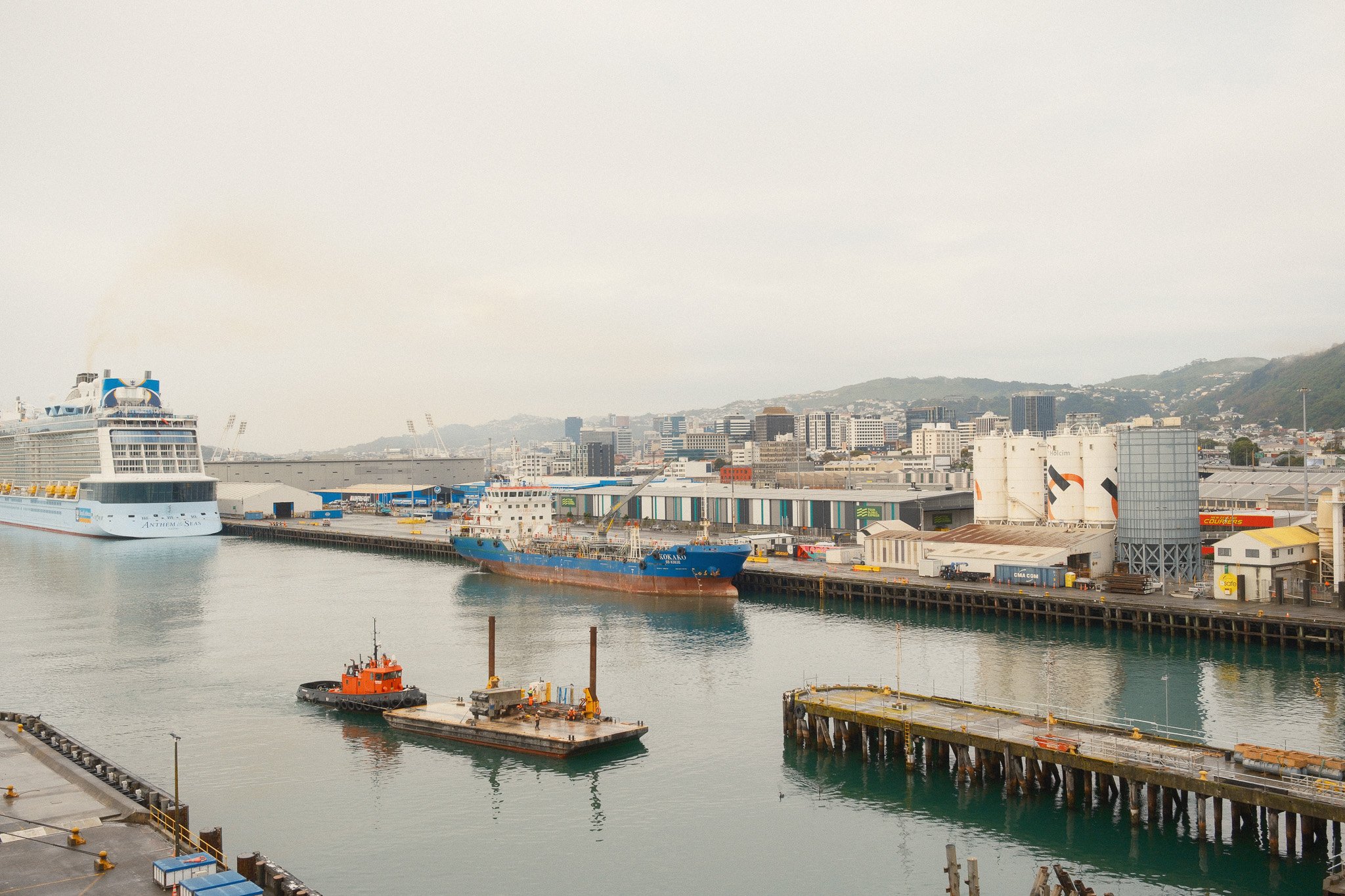 A harbor scene with a large cruise ship and a smaller cargo ship docked along the pier, with industrial warehouses and city buildings in the background under an overcast sky.