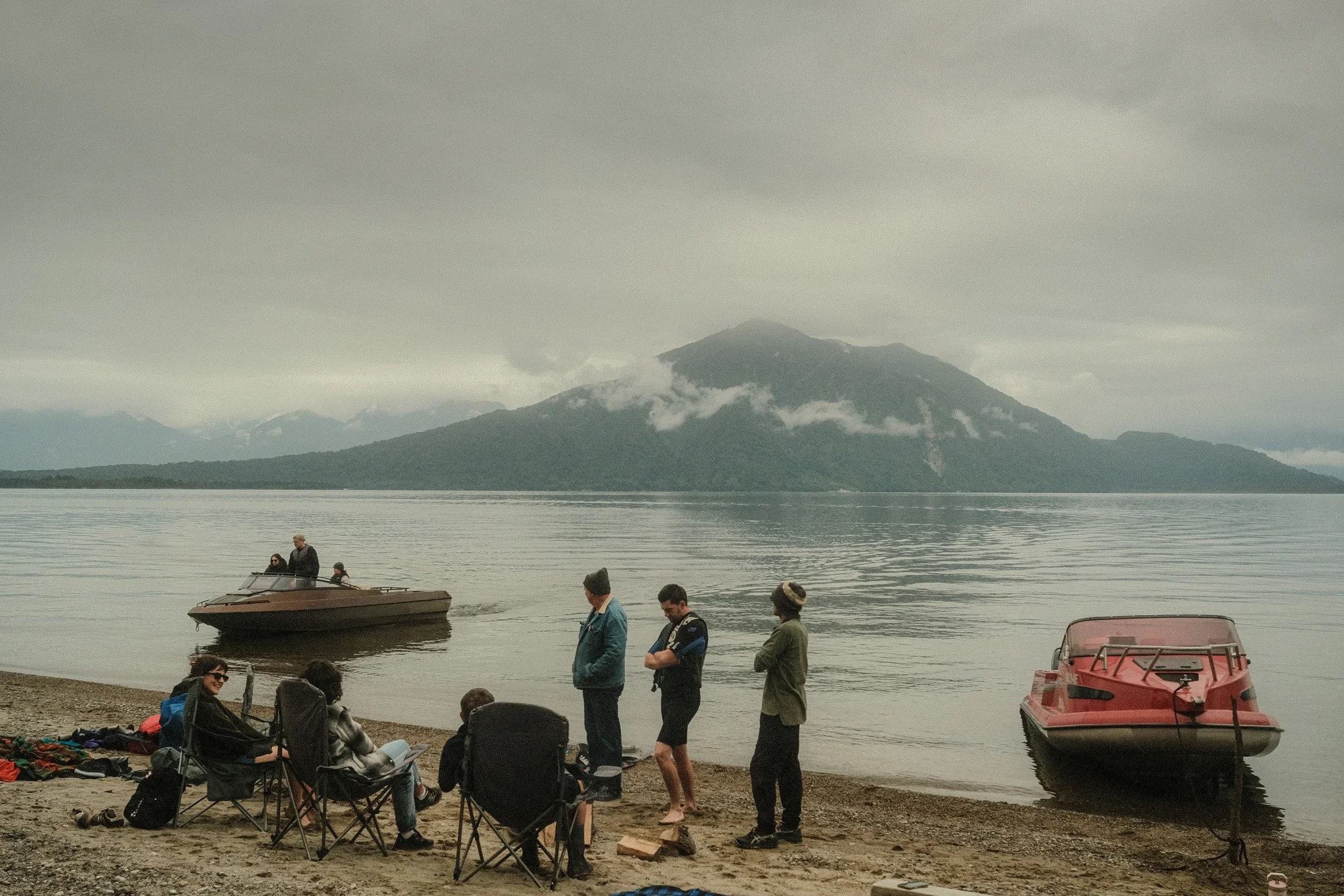 People on a beach with two boats, and a mountain and cloudy sky in the background.