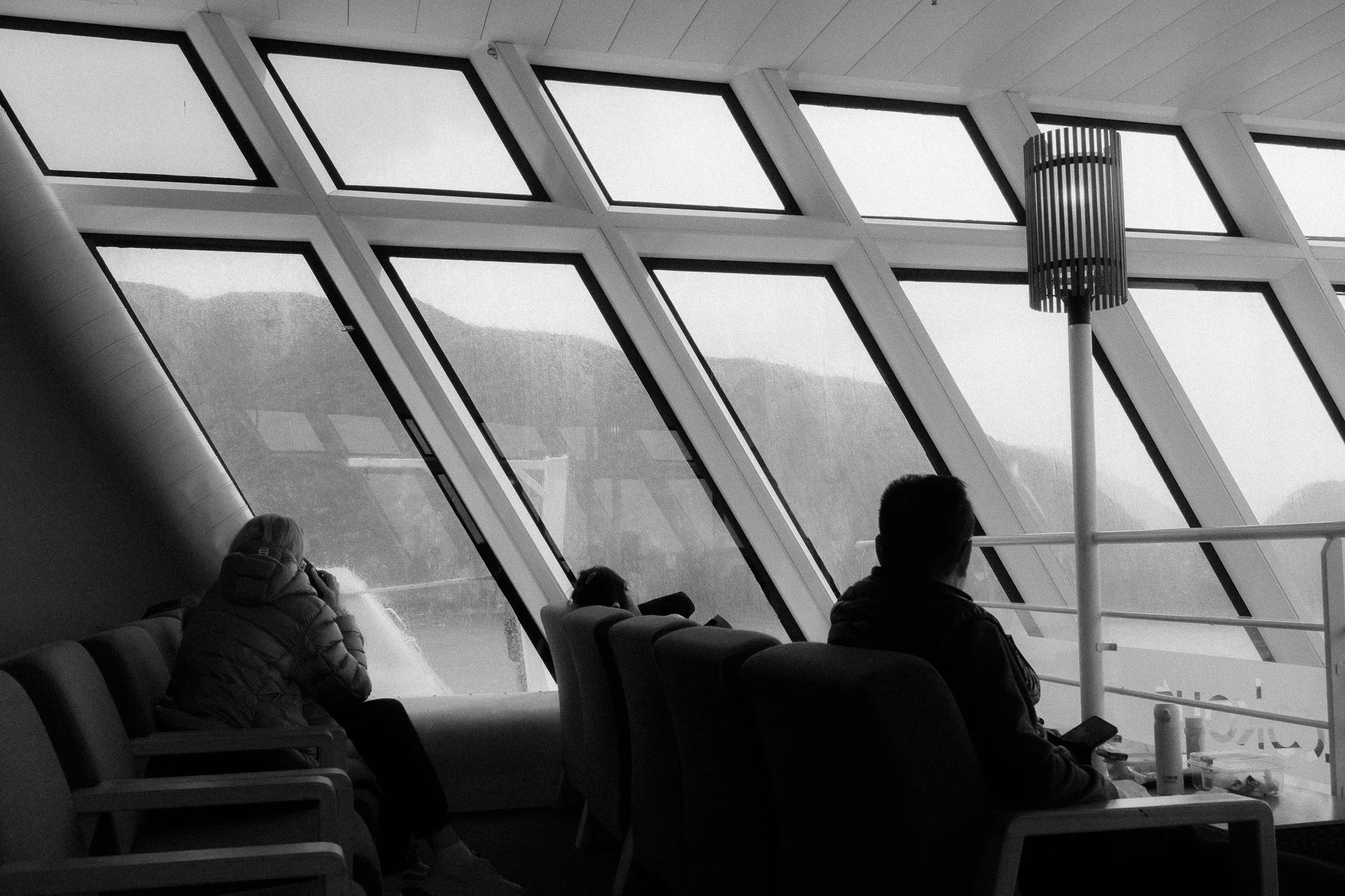 People sitting inside a modern building with large, angled windows revealing an outdoor landscape with hills, a person on the left taking photos, and others using mobile devices.