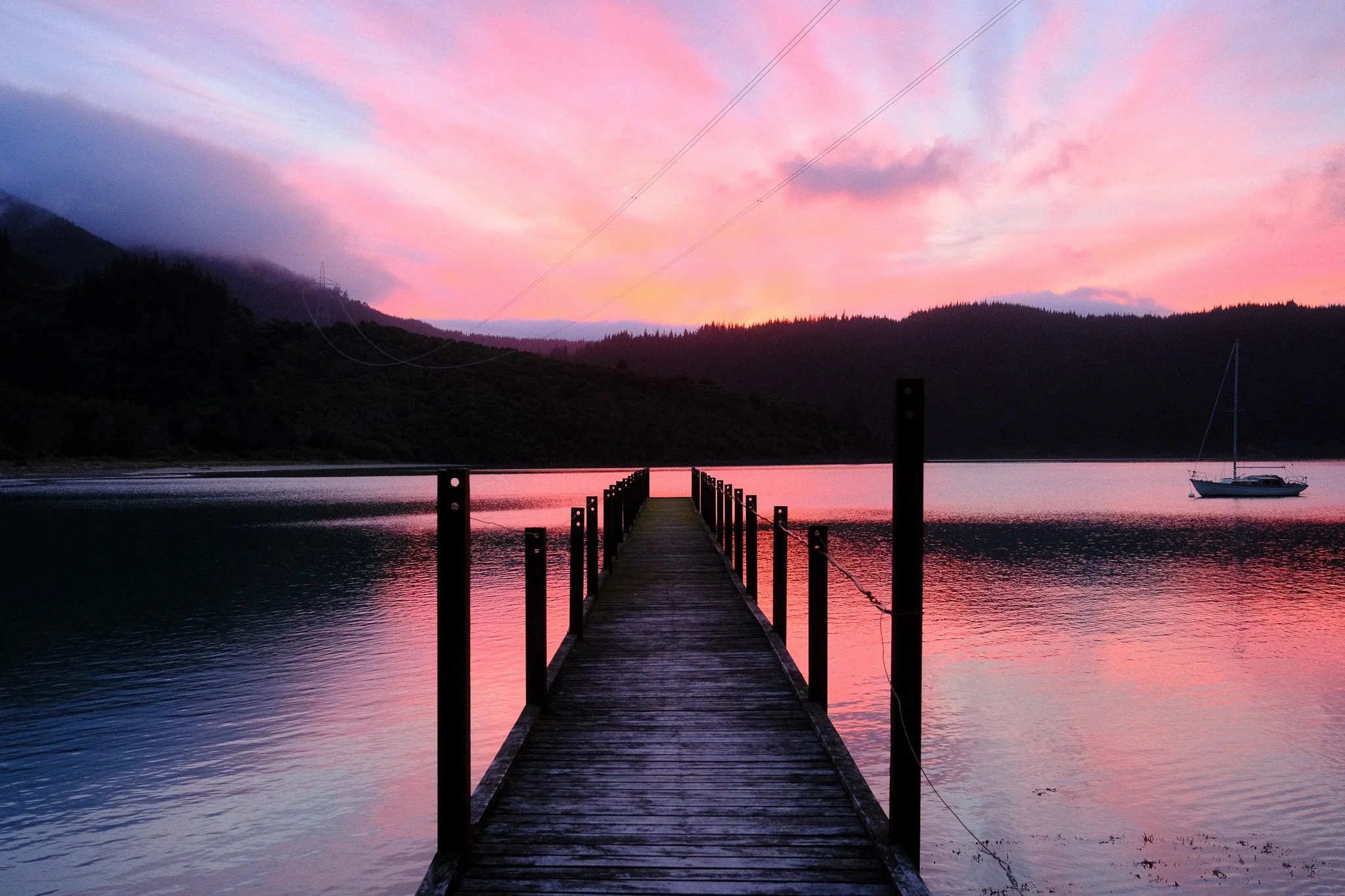 A wooden dock extends into a calm lake during sunset, with pink and purple hues in the sky and a sailboat anchored on the right side of the water.
