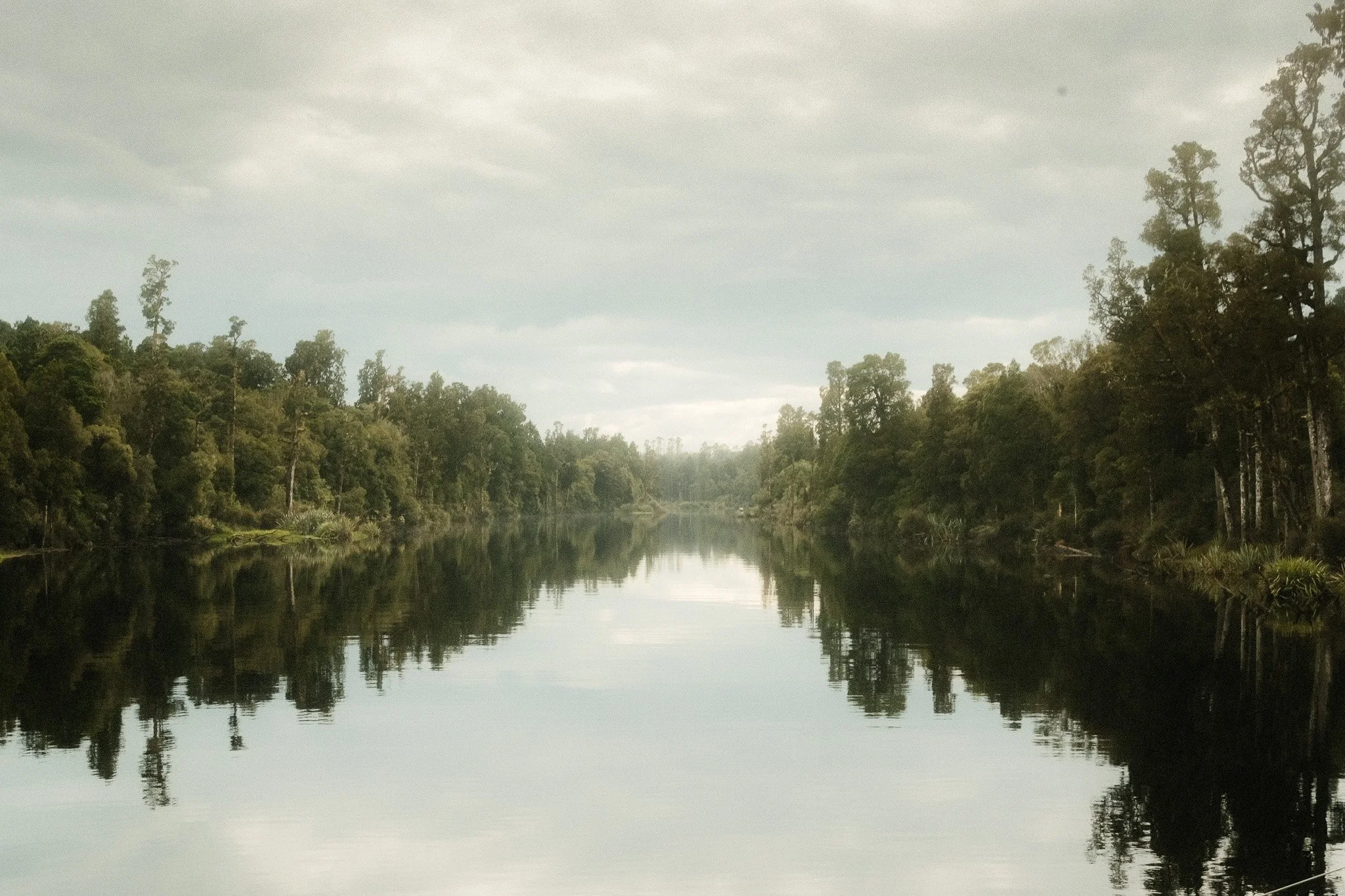 A calm river reflecting dense trees on both sides under a cloudy sky.