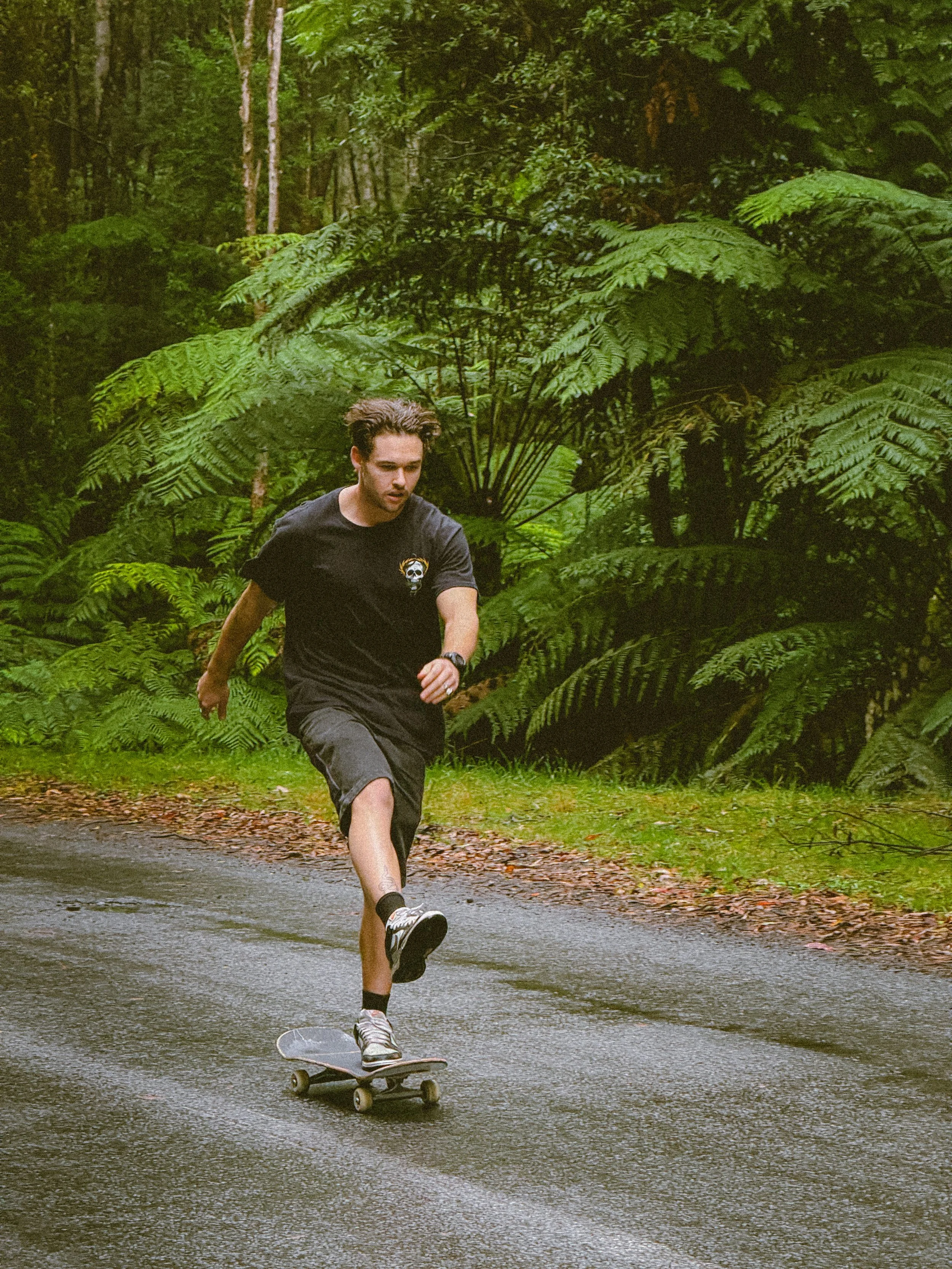 A young man skateboarding on a wet paved road through a dense green forest.