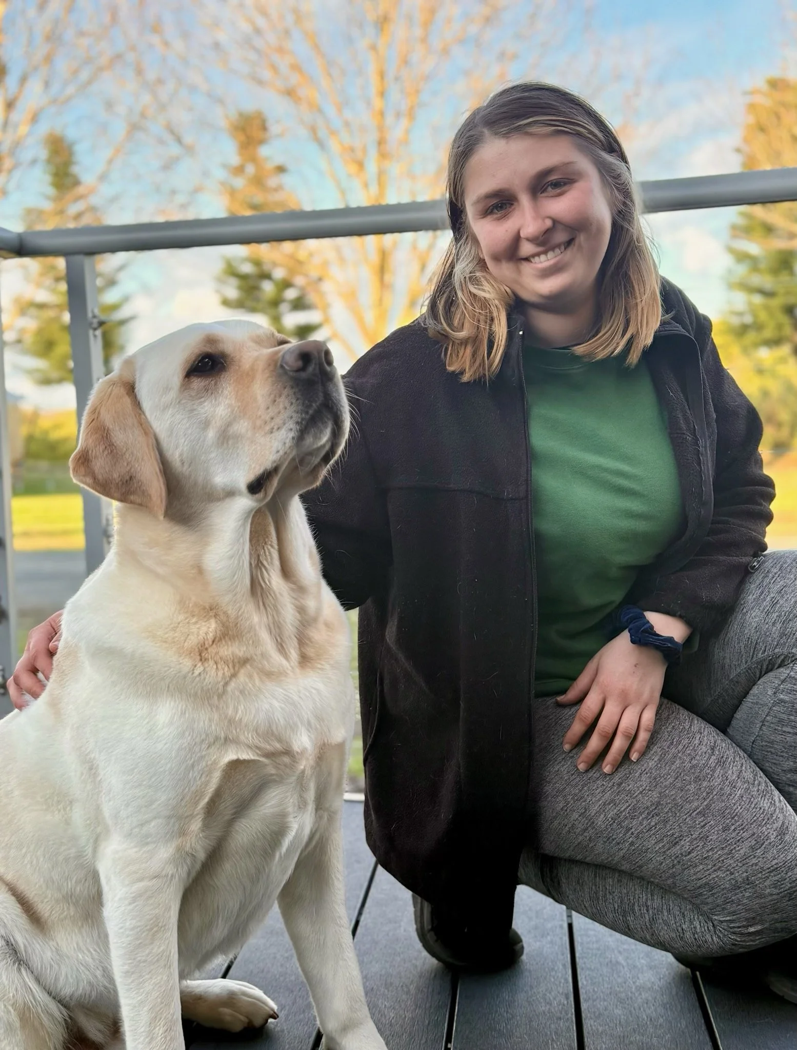 A young woman kneeling on a deck with her arm around a yellow Labrador retriever dog. She has shoulder-length blonde hair, a black jacket, a green shirt, and gray leggings. The dog is sitting, looking attentive. The background features trees with autumn foliage and a clear sky.
