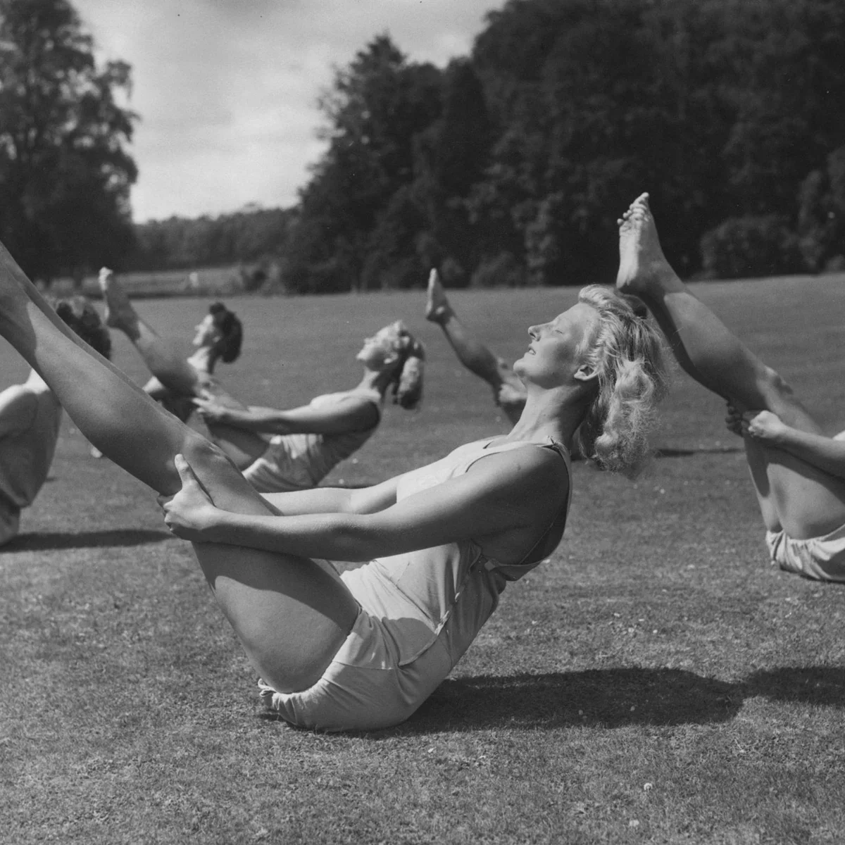 A group of women in vintage summer clothing practices yoga outdoors on a grassy field on a sunny day, with trees in the background, in black and white.