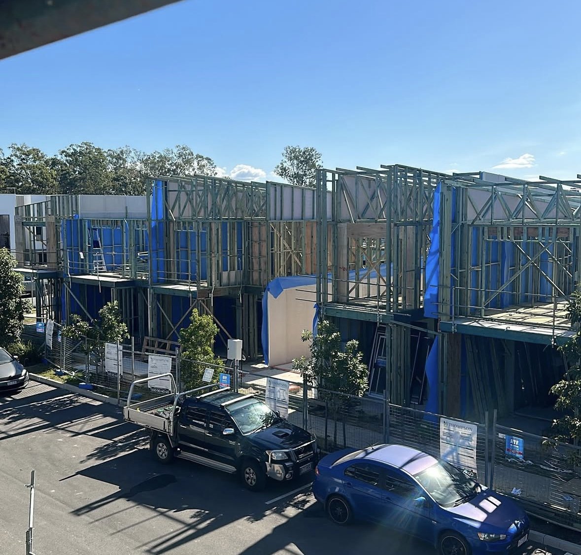 Construction site with a building framework of metal studs and blue sheathing, parked cars in front, and a clear blue sky.