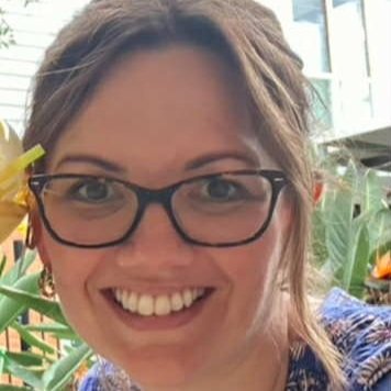 Close-up of a woman with glasses, smiling, outdoors among green plants and flowers.