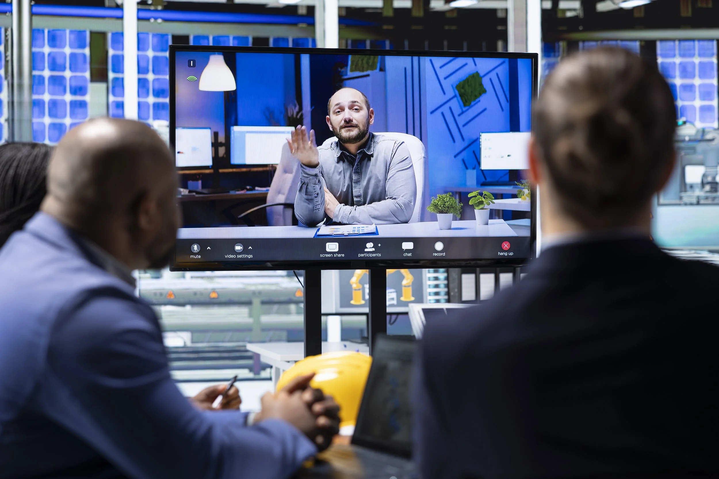 Three business professionals in a meeting room watching a video conference on a large screen. The screen displays a man with a beard and a gray shirt speaking to them from an office.