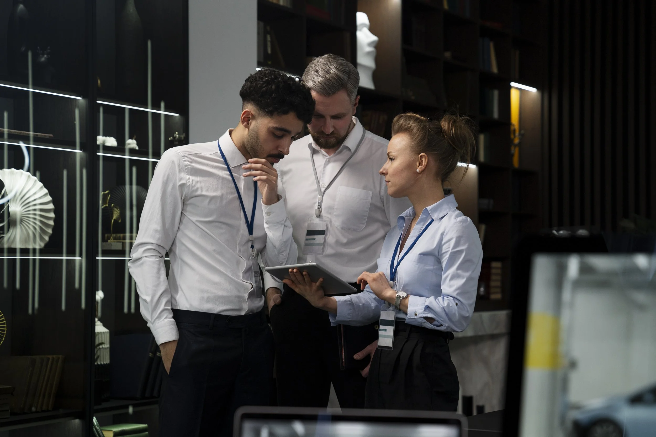 Three people, two men and one woman, standing together in business attire, looking at a tablet device in a modern office or conference room.
