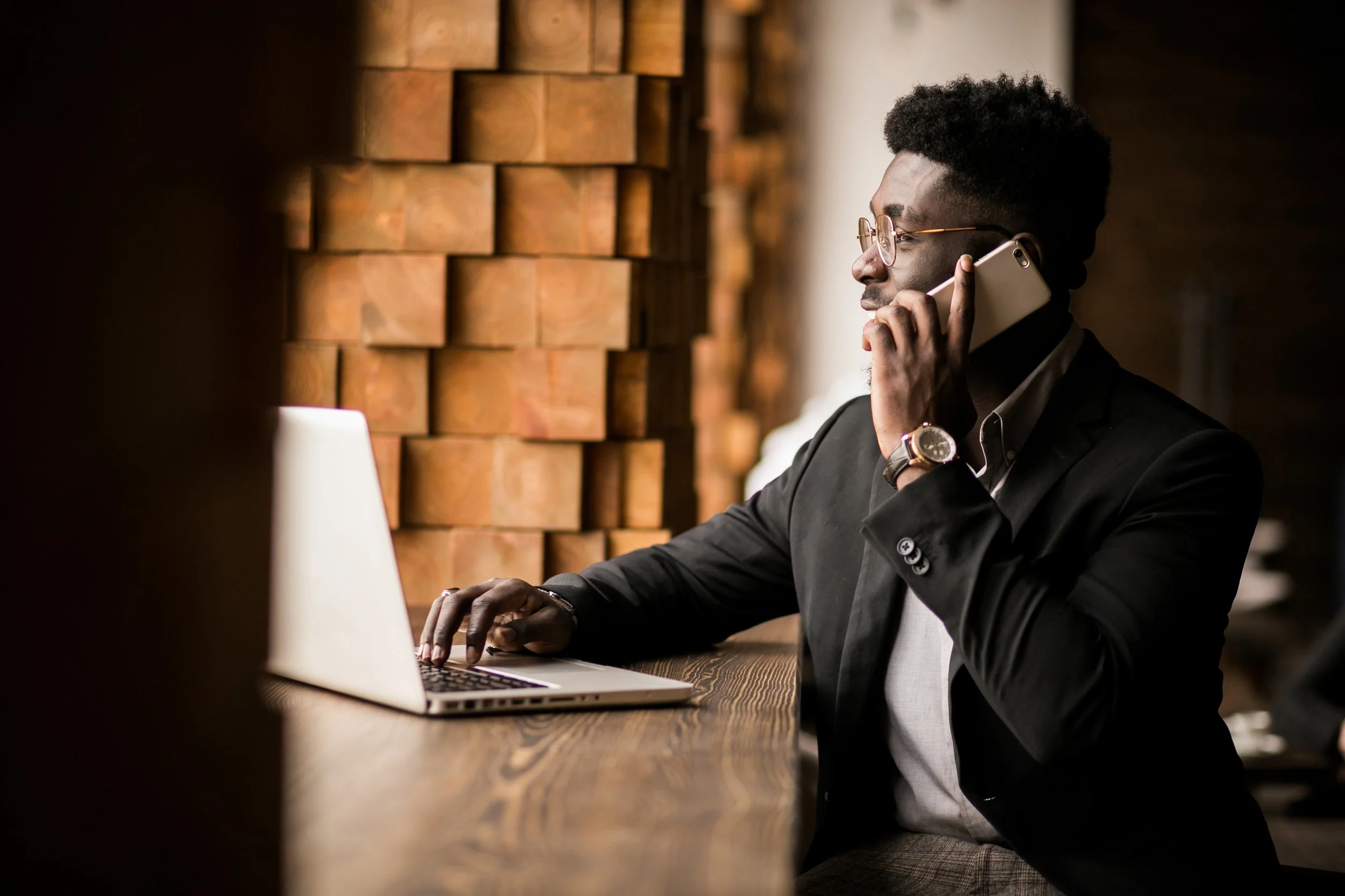 A man in a black suit and glasses sitting at a wooden table, talking on a cellphone and working on a silver laptop in a modern setting with a brick accent wall.