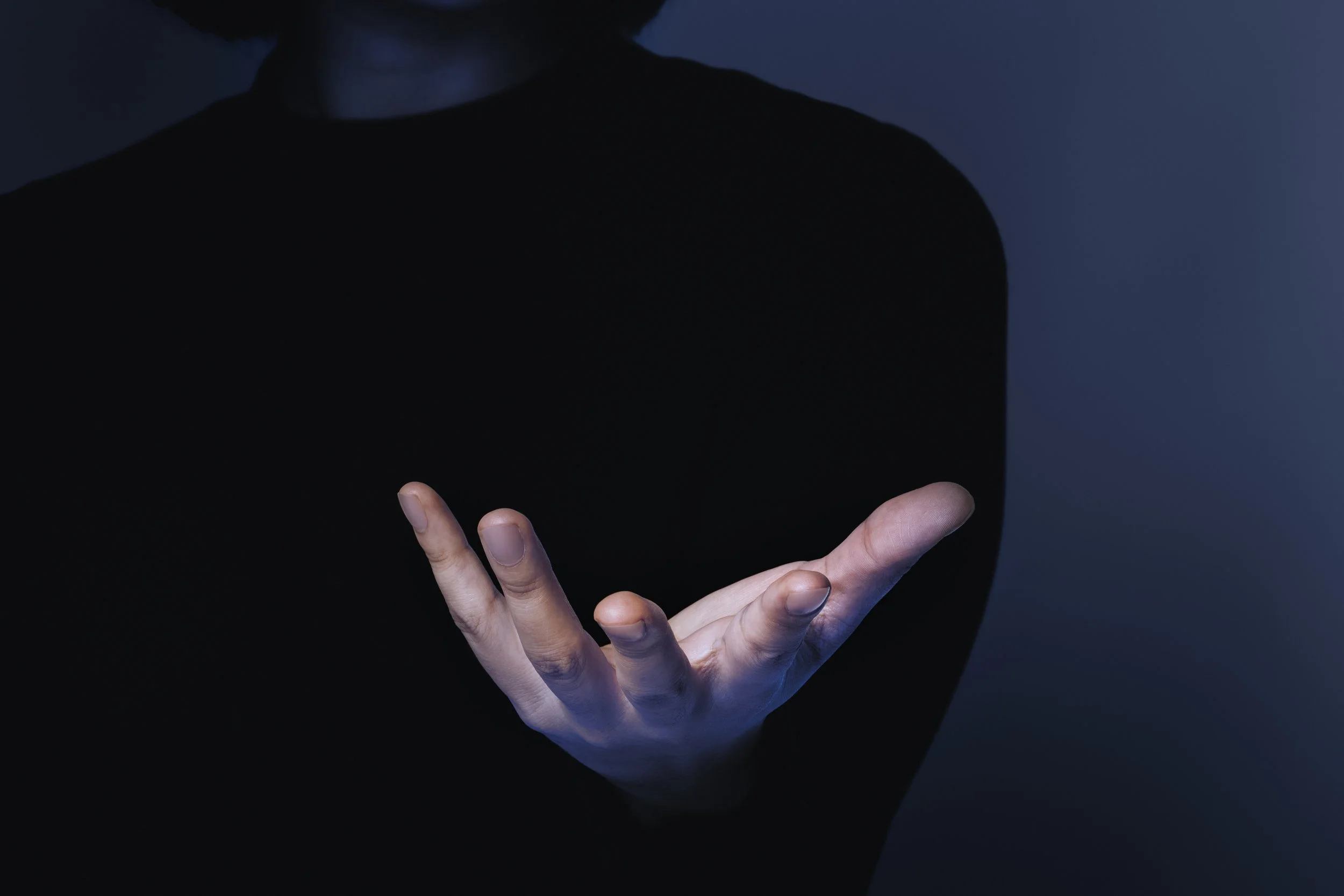 A person in a black shirt extending their hand forward with palm up, set against a dark background.