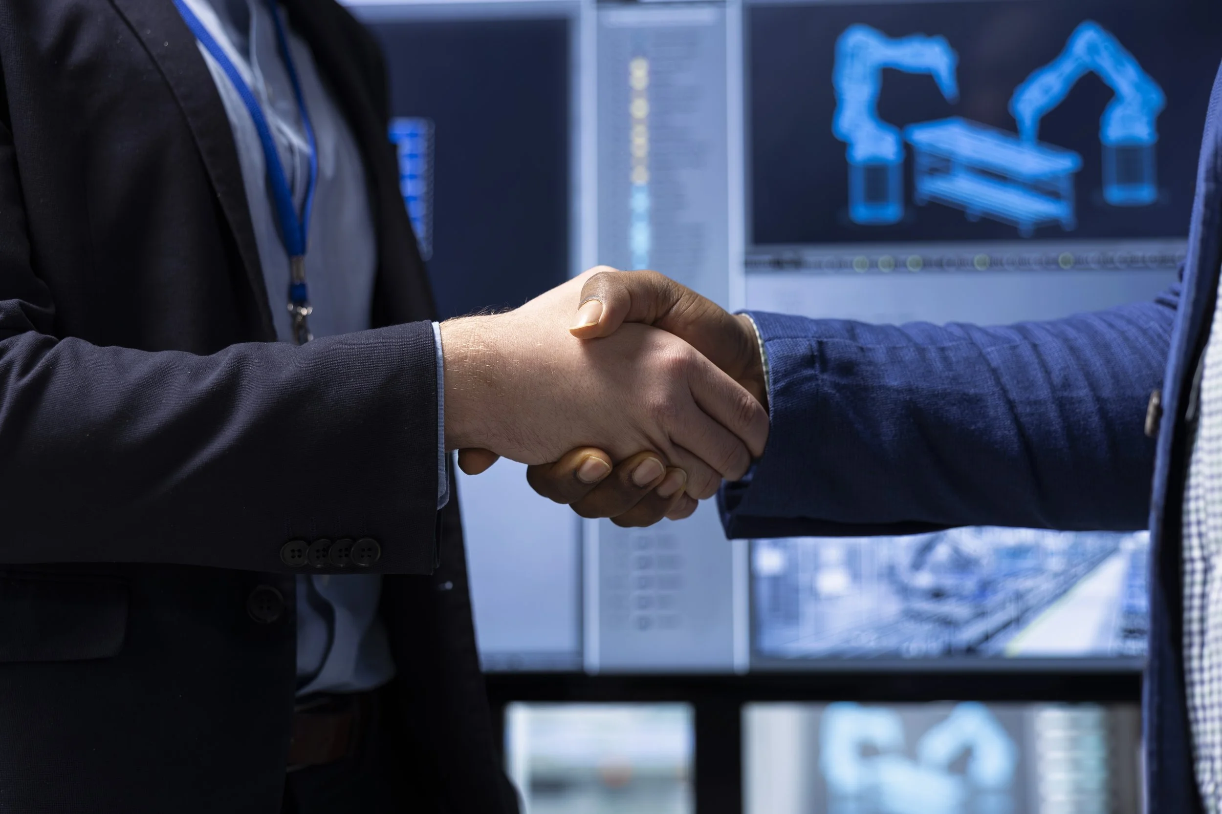 Two people in business suits shaking hands in front of computer monitors displaying architectural or engineering designs.