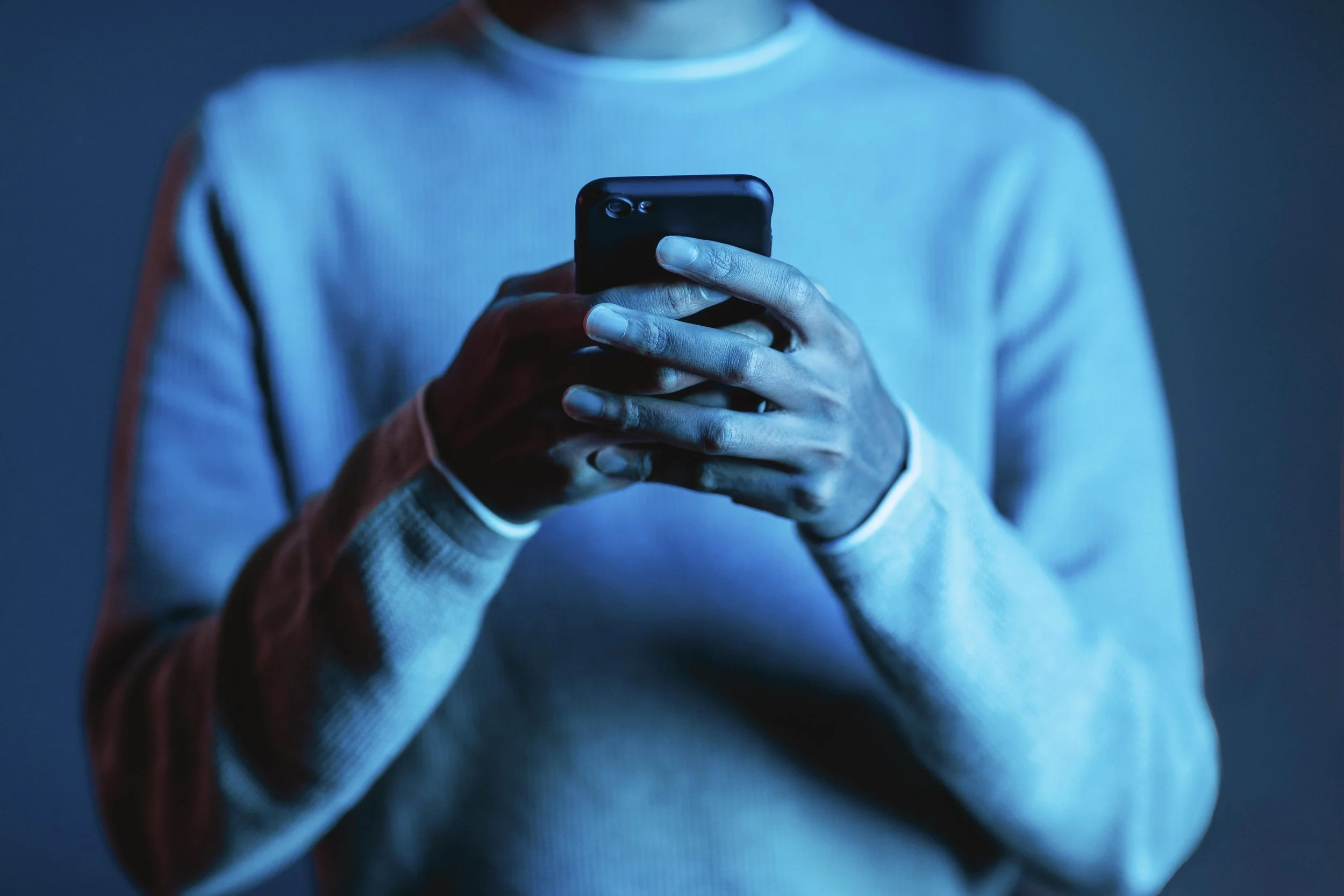 Person in a gray sweater looking at a smartphone in a dark room with blue lighting.