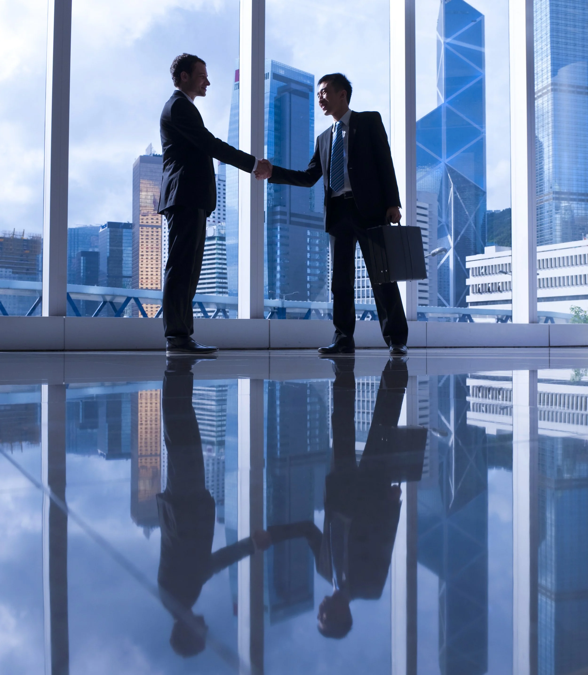 Two businessmen shaking hands in a modern office with a city skyline view, one carrying a briefcase.