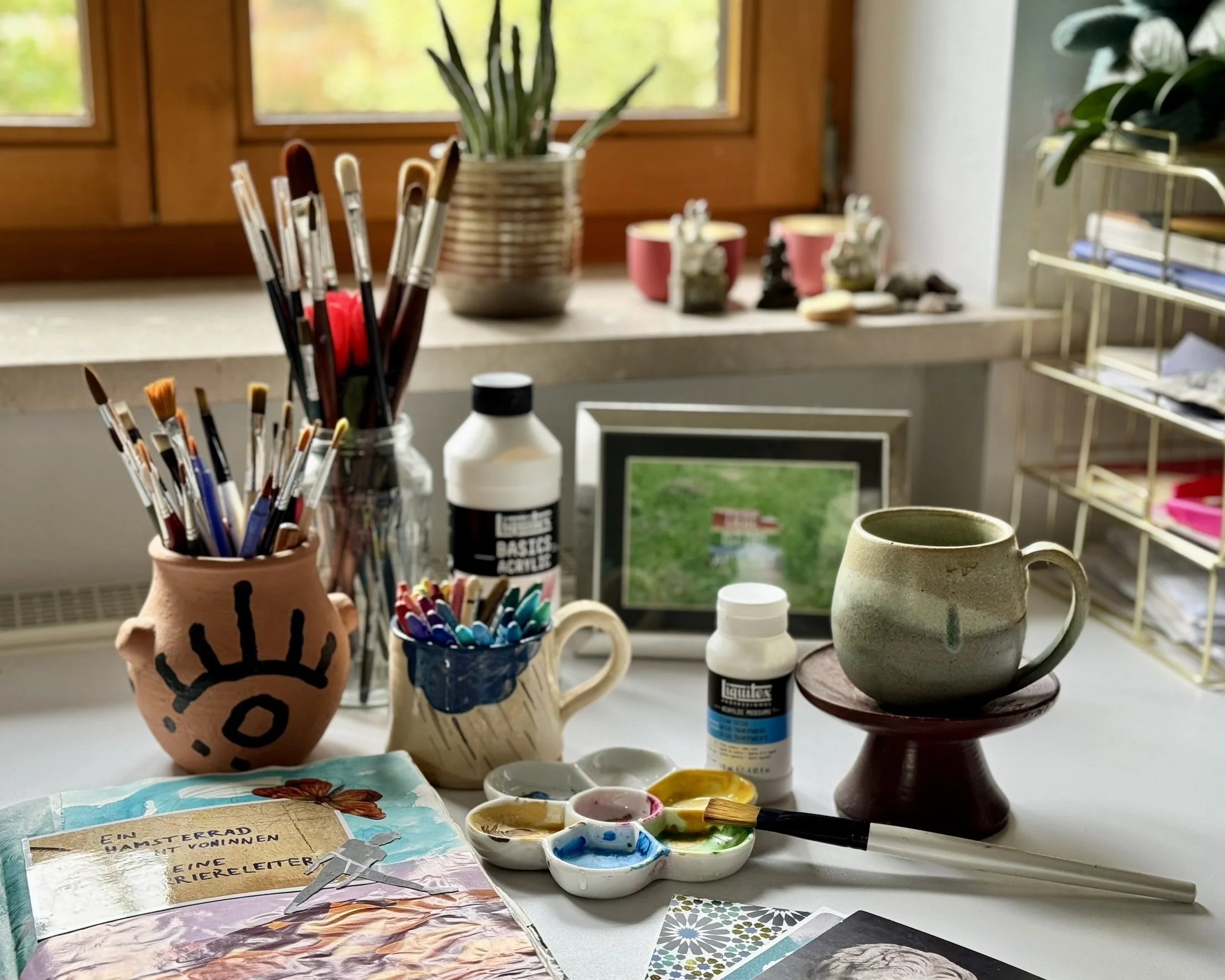 Ein Arbeitsplatz zum Malen und Basteln mit Farbpalette, Pinsel, Keramikbechern, Fotografien, Pflanzen und Bastelmaterialien auf einer weißen Arbeitsfläche vor einem Fenster.