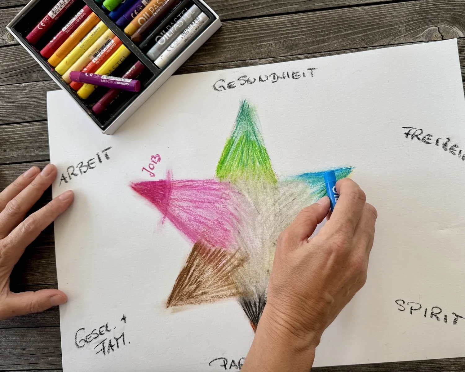 Auf einem weißen Blatt Papier ist ein bunter, sechszackiger Stern gemalt. Man sieht, wie die linke Hand das Papier festhält, die rechte Hand mit hellblauer Ölkreide das Bild bearbeitet.