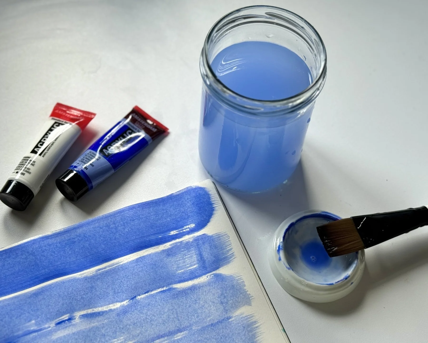 Kunstmaterialien auf einem weißen Tisch, darunter ein Glas mit blauer Farbe, zwei Tuben Acrylfarbe, ein Pinsel in einer kleinen Schale mit blauer Farbe und eine Skizzenbuchseite mit blauen Pinselstrichen.