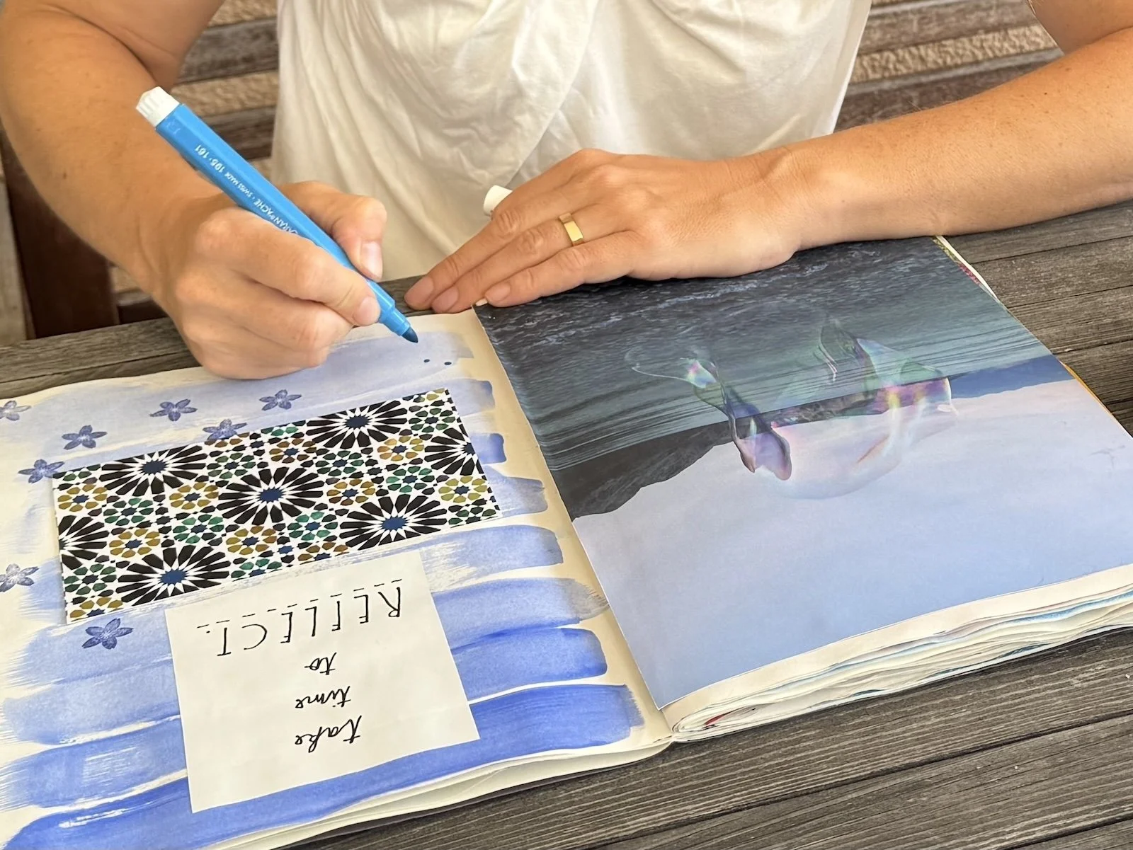 Eine Person schreibt mit einem blauen Marker in ein aufgeschlagenes Buch. Auf der linken Seite ist eine collageartige Seite mit Mustern und einem handgeschriebenen Text, während auf der rechten Seite eine Fotografie eines Wasserfalls und eines Sees z