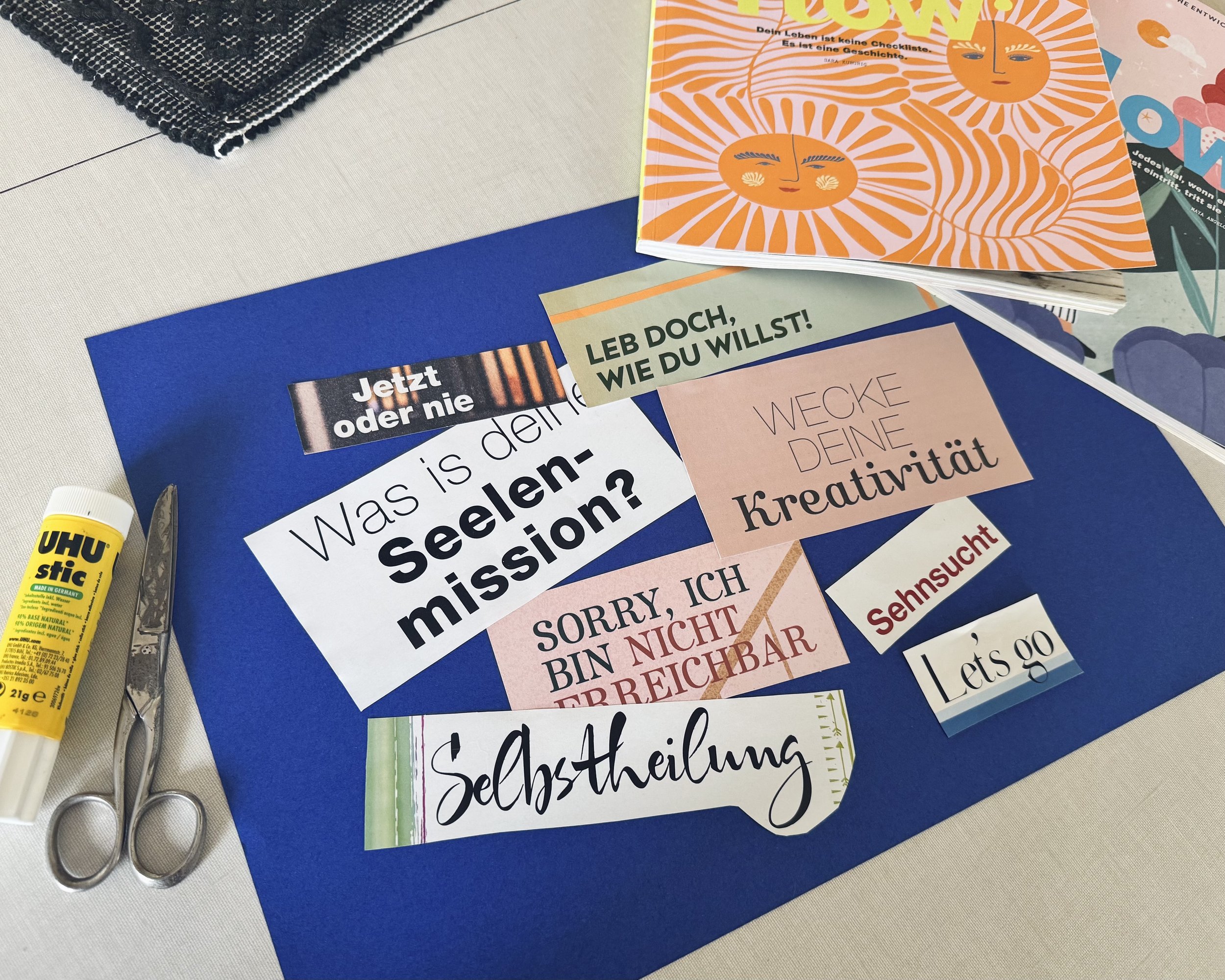 Auf einem königsblauen Fotokarton liegen verschiedene Ausschnitte aus Zeitschriften, größtenteils Worte wie "Was ist deine Seelenmission?" oder "Selbstheilung". Eine Schere und Uhu-Kleber liegen daneben, ebenso zwei bunte Zeitschriften