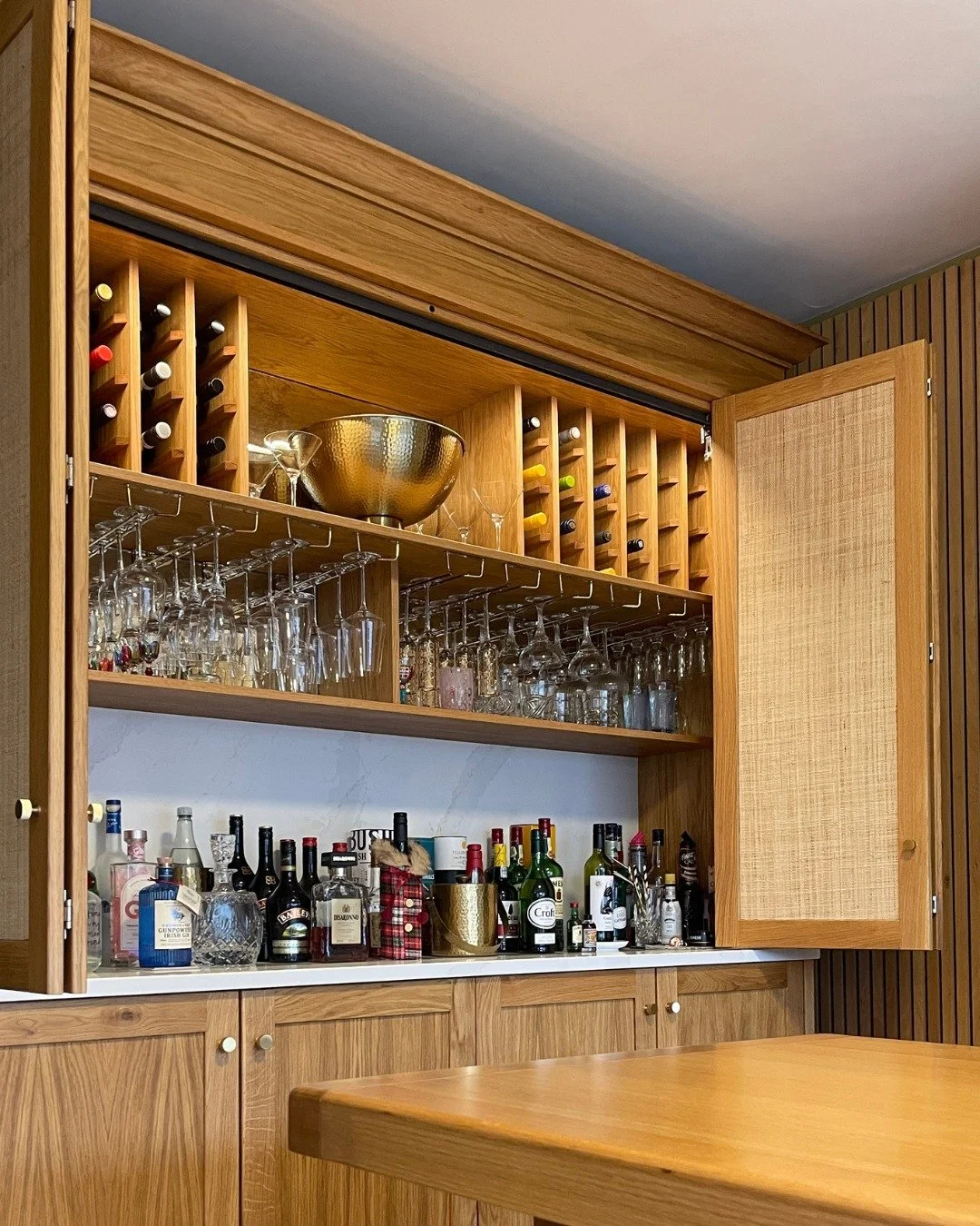 Evenings often begin here without much thought. Antique mirrors add character, while the Calcutta Gold stone worktop offers a surface for mixing and serving drinks. Woven raffia panels paired with oak bring warmth and texture, keeping the space relax