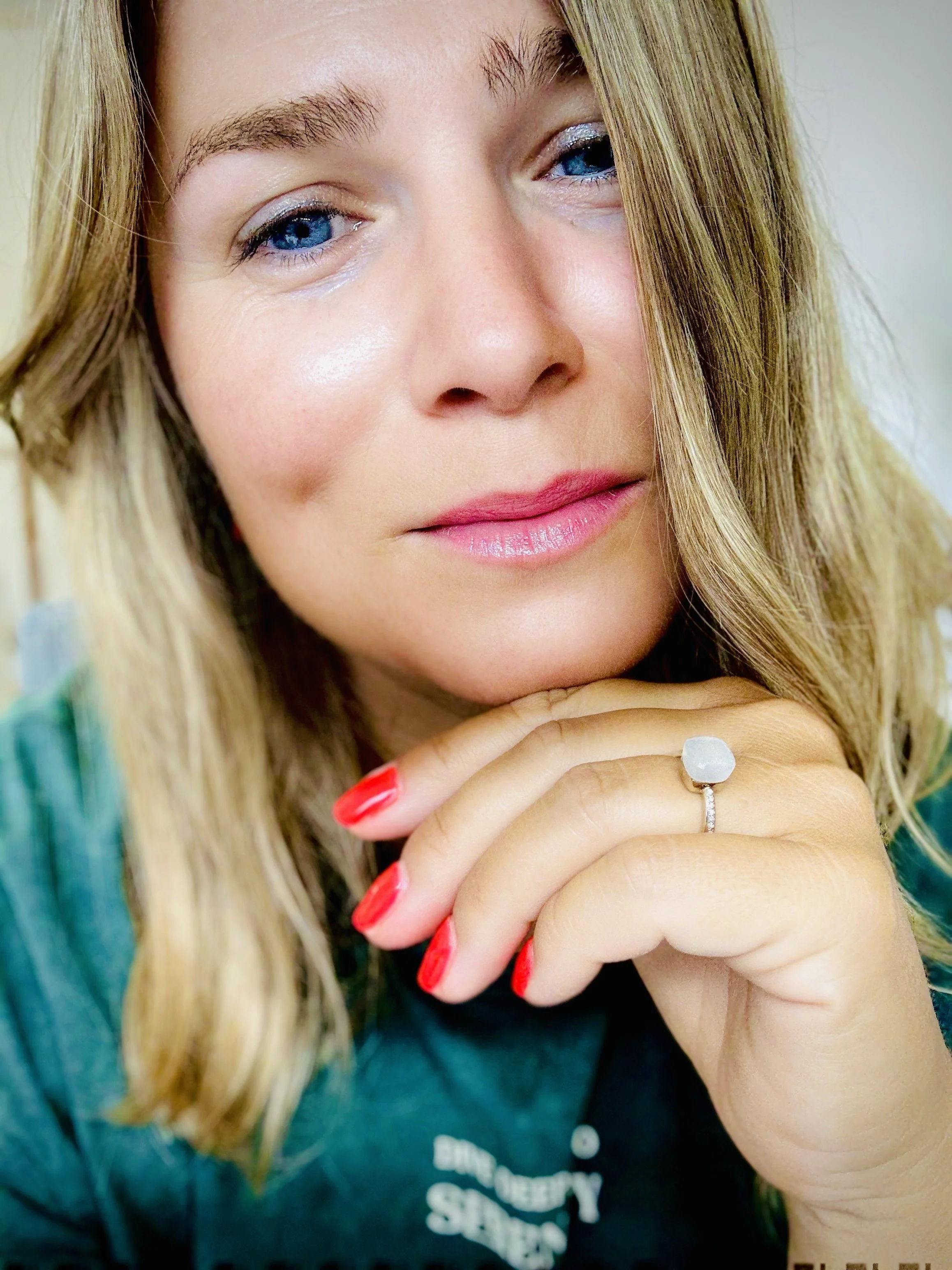 Zeljka Cacic-Escalera, a Tony Robbins and Chloe Madanes Trained Coach, with blonde hair, blue eyes, pink lipstick, and red nail polish, resting her chin on her hand with a ring featuring a large, light-blue gemstone.