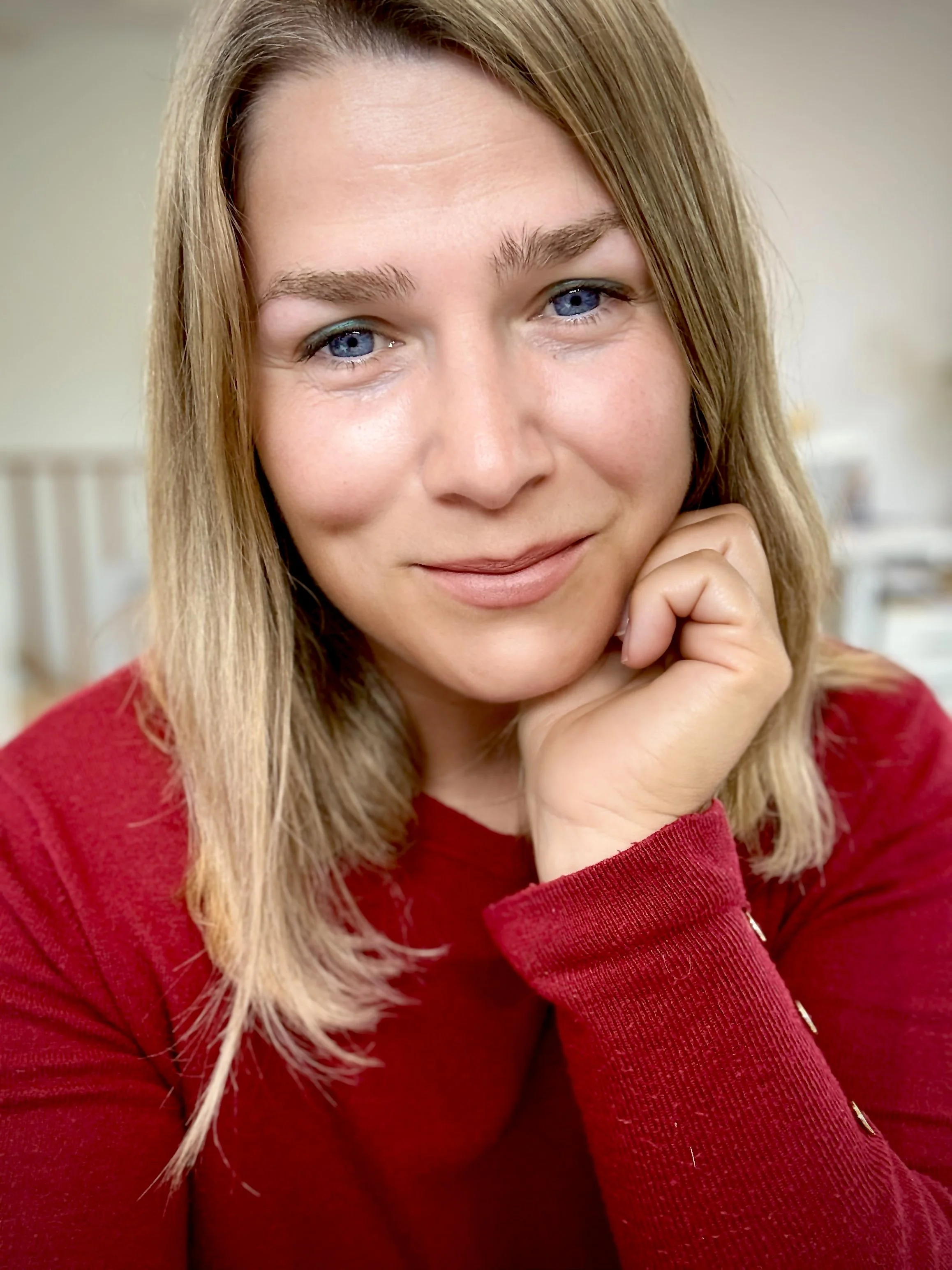Zeljka Cacic-Escalera, a Tony Robbins and Chloe Madanes Trained Coach, with blonde hair and blue eyes, wearing a red sweater, resting her chin on her hand and smiling softly.