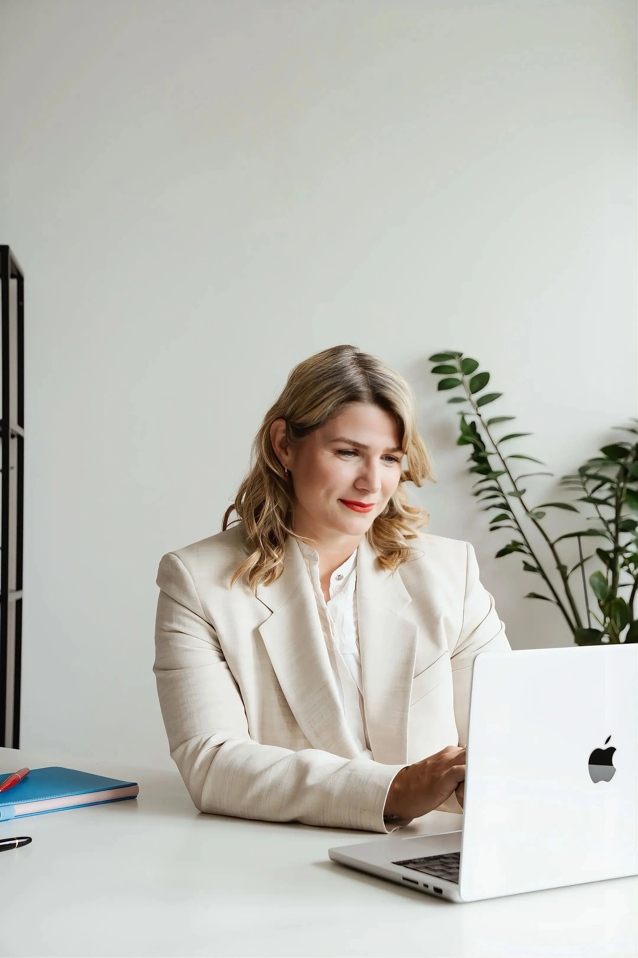 An image of Zeljka Cacic-Escalera, a GLP-1 and Transformation Coach in Oceanside, California. She is wearing a beige blazer working on a laptop at a desk with a blue notebook and a red pen, with a green plant in the background.