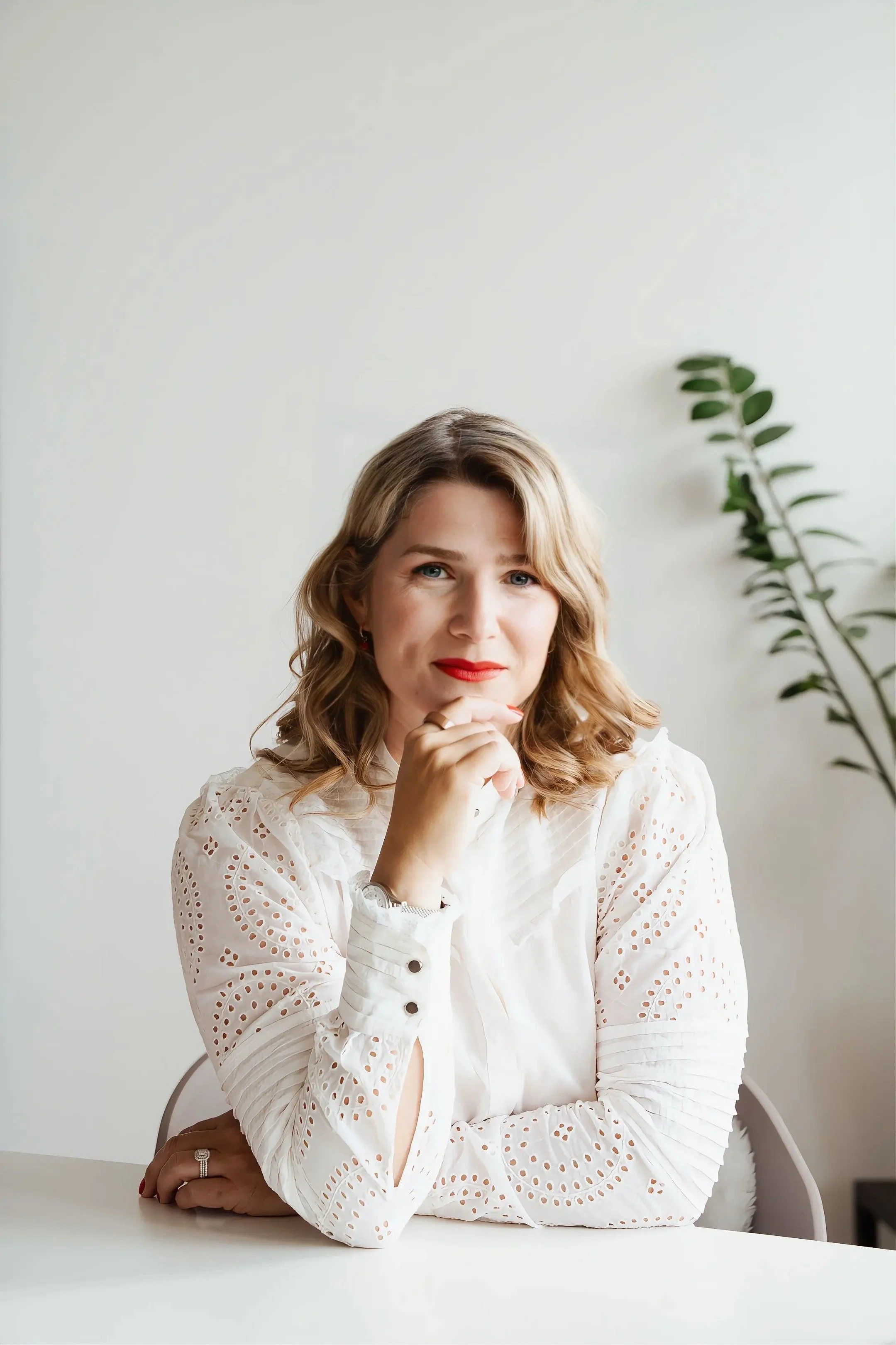 An image of Zeljka Cacic-Escalera, a GLP-1 and Transformation Coach in Oceanside, California. She has red lipstick and blue eyes sitting at a white table, resting her chin on her clasped hands, with a green plant in the background.