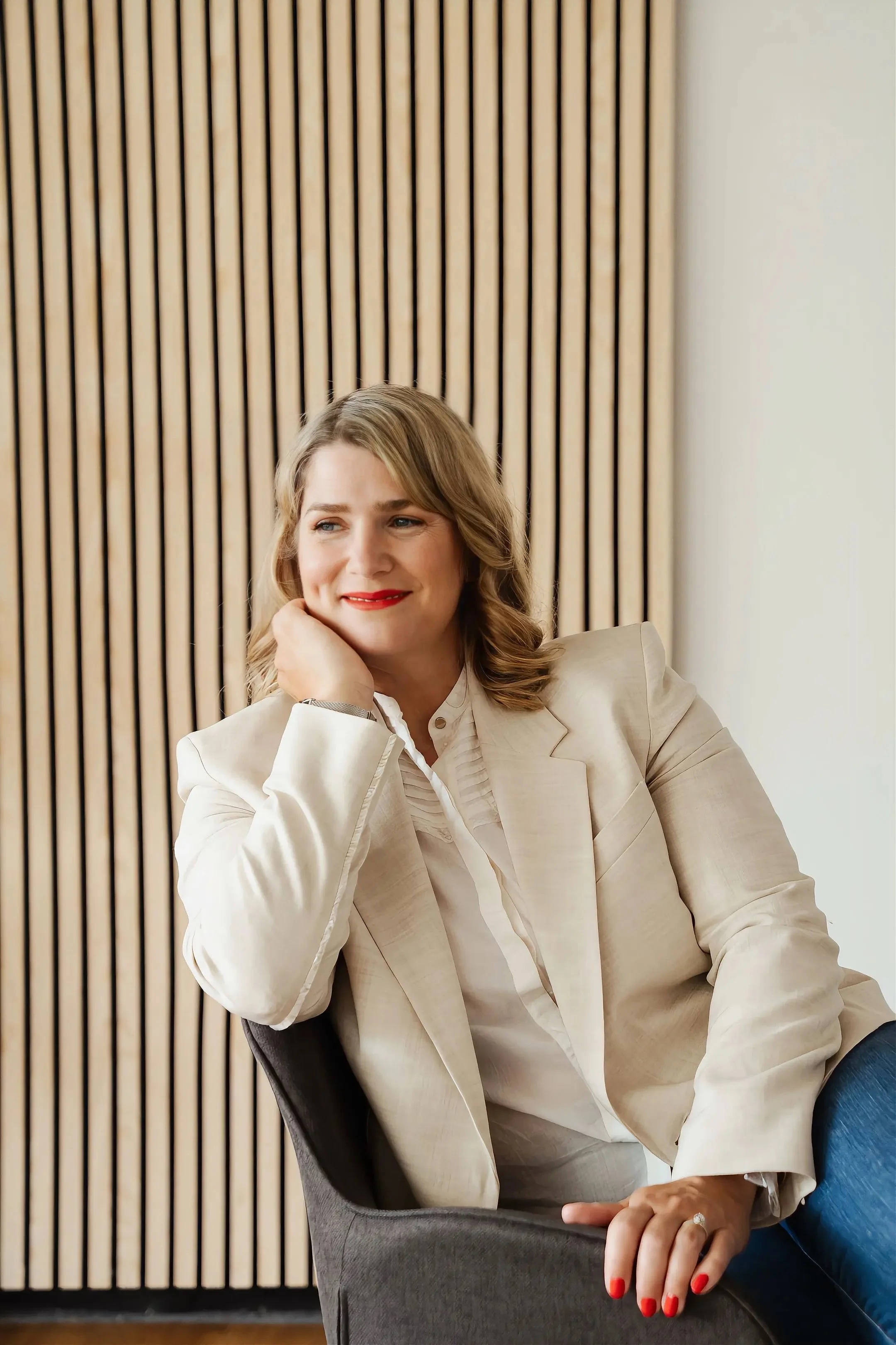 An image of Zeljka Cacic-Escalera, a GLP-1 and Transformation Coach in Oceanside, California. She has blonde hair and red lipstick as is sitting in a chair, wearing a beige blazer and white shirt, with nail polish and a ring on her finger. 